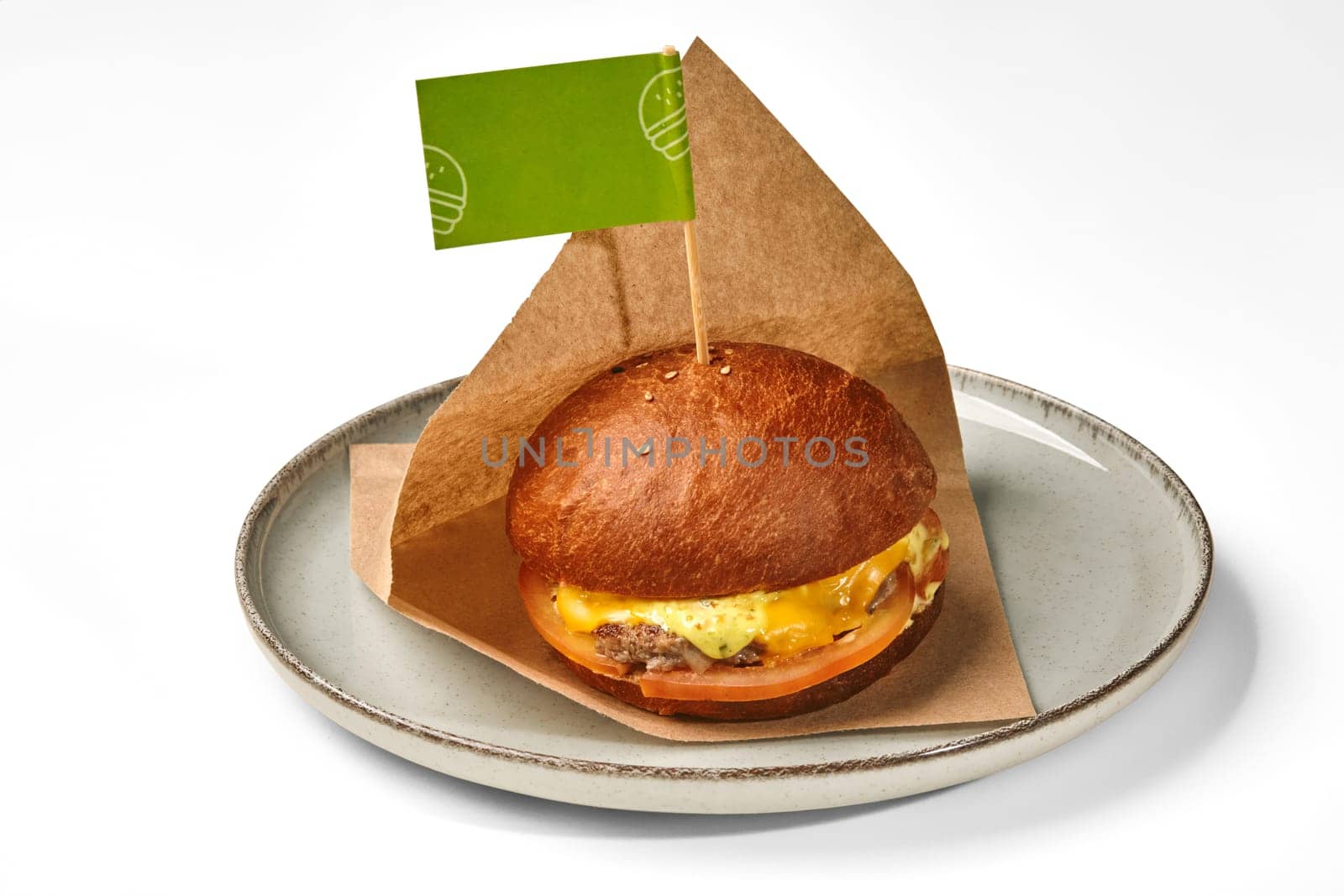 Appetizing hamburger in toasted fluffy bun with juicy beef patty, tomatoes and spicy cheese sauce, served in paper wrap with green flag label on plate against white background