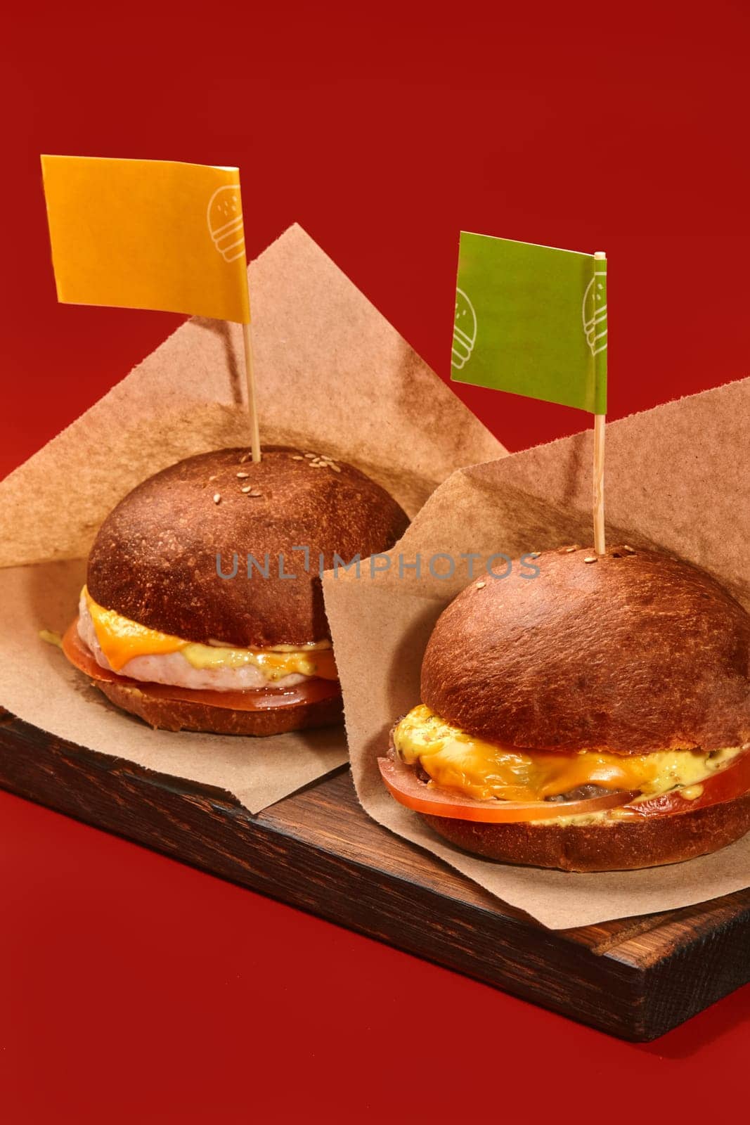 Two delicious burgers with juicy chicken and beef patties, tomato and cheese in browned buns decorated with colorful flags on toothpicks, served in craft paper on wooden serving board on red backdrop