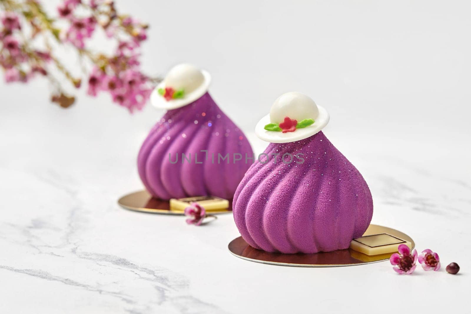 Chic velvety purple fig-shaped mousse confections topped by white chocolate hats, styled with delicate spring florals on marble table. Collections of signature handmade desserts