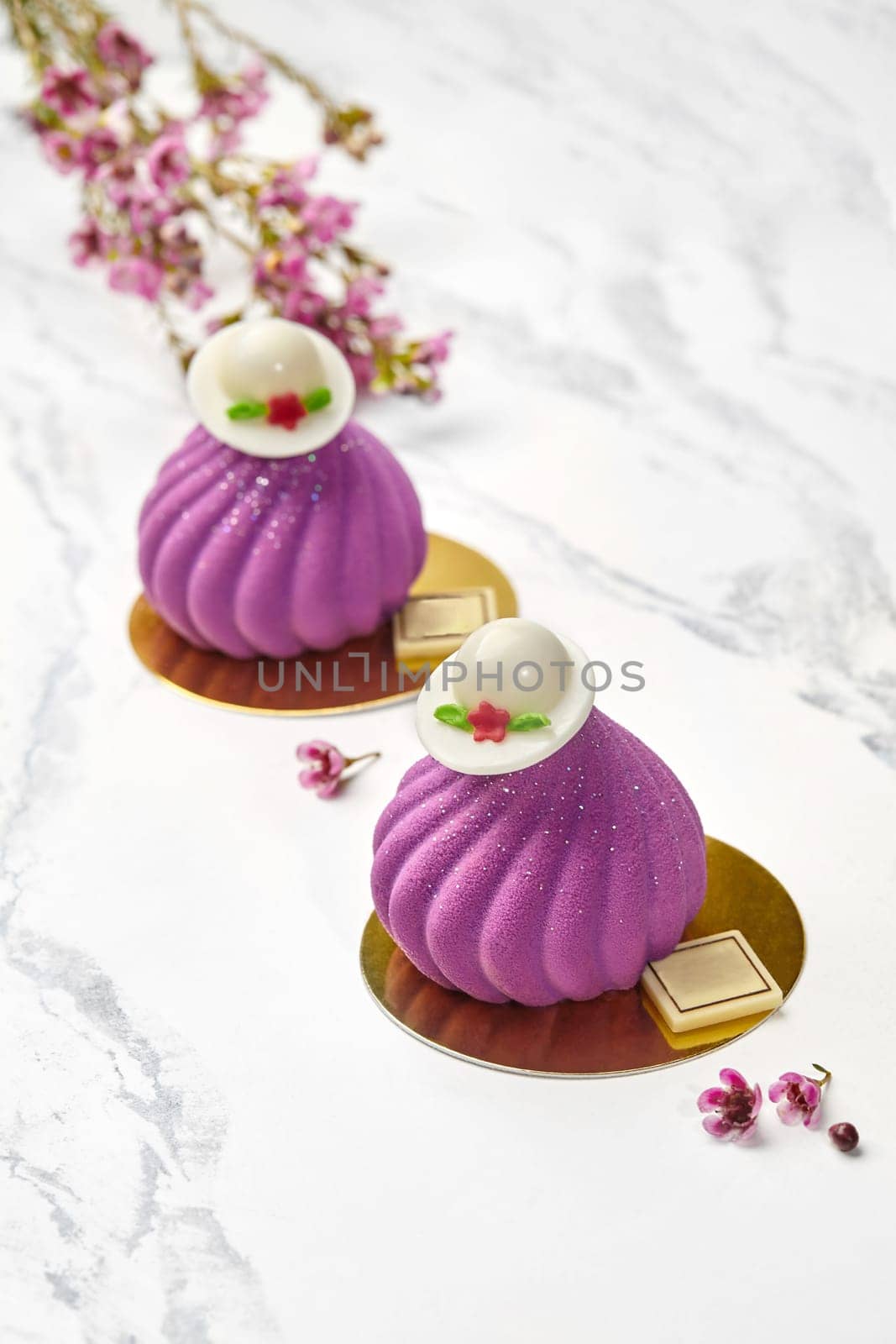 Sophisticated mousse pastries in vibrant purple with velvet texture, crowned with decorative white chocolate caps, set against backdrop of pink blossoms and marble