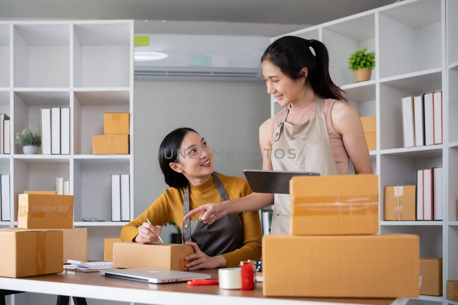 Couple of young Asian women running a small business together selling products online using laptops by wichayada