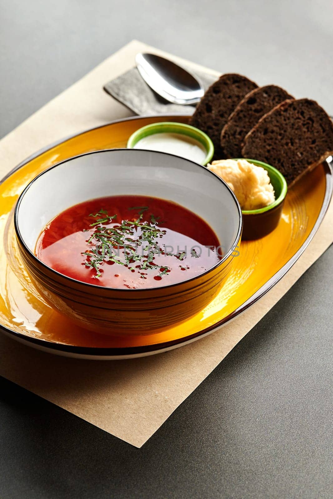 Elegant presentation of classic borscht soup sprinkled with chopped greens served on yellow tray and paper placemat with dark rye bread, sour cream, and condiments. Traditional Ukrainian dish