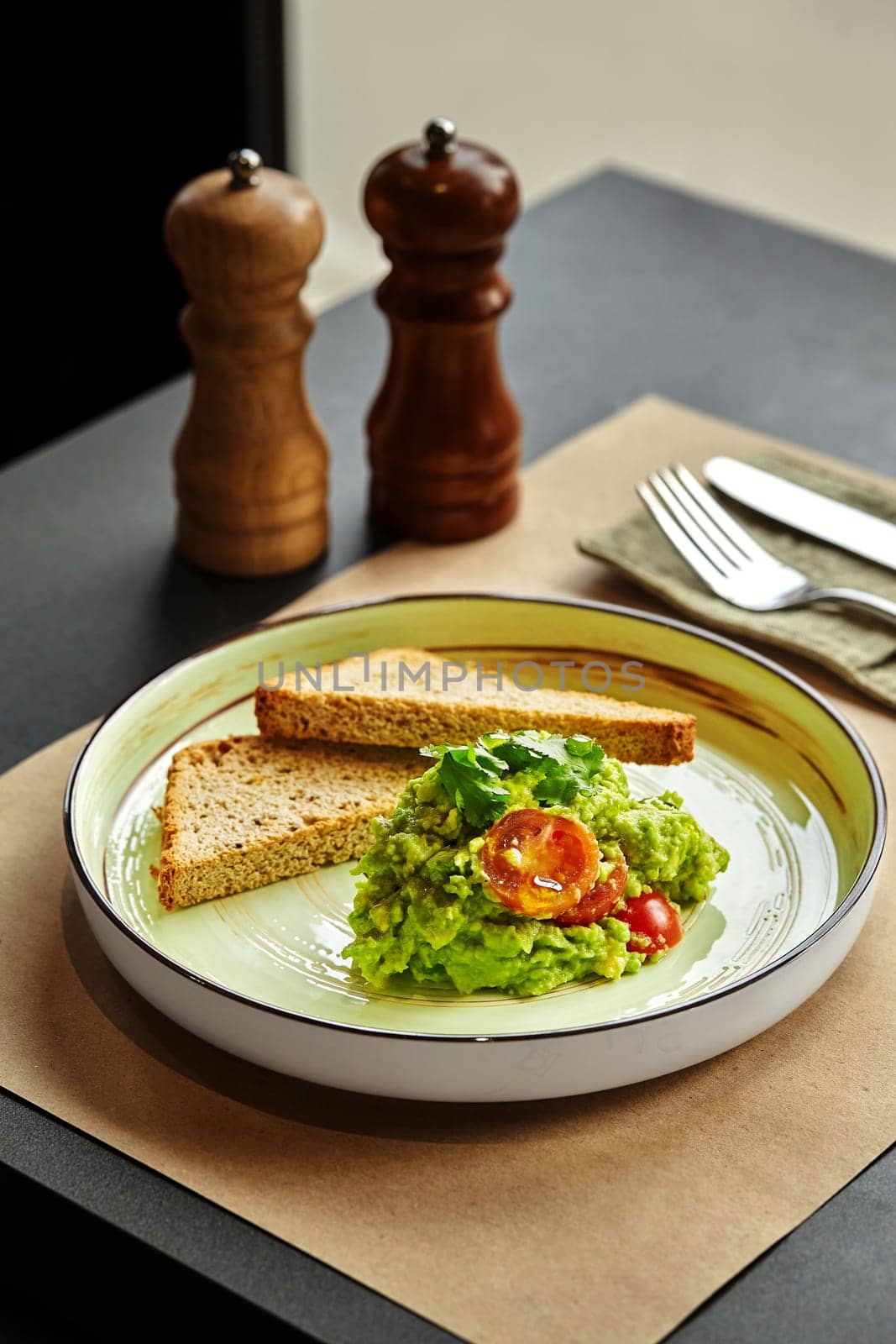 Vibrant freshly mashed piquant guacamole with juicy cherry tomatoes and green herbs served on plate with crispy toasts, accompanied by wooden salt and pepper shakers. Light Mexican style snack concept