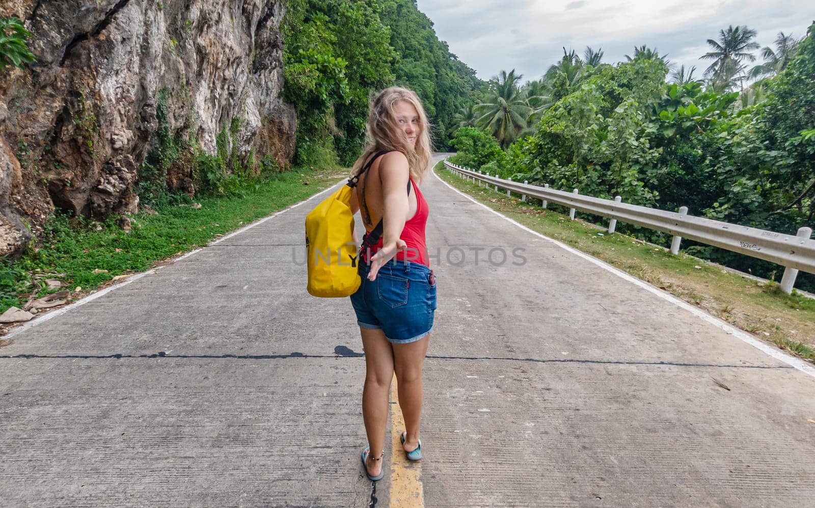 A young woman with a yellow backpack appears to be inviting the viewer to join her as she explores a deserted road lined with dense, green foliage and towering cliffs.