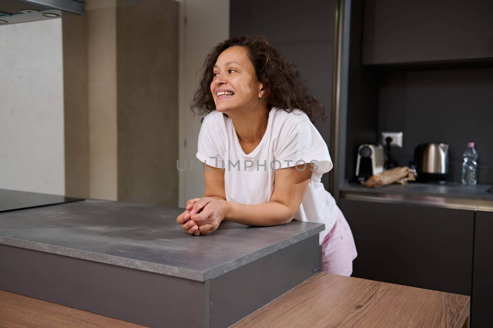 Beautiful curly brunette woman in pajamas, smiling with cheerful toothy smile, looking aside while standing at kitchen counter,enjoying a happy morning of her holidays at home. People. Lifestyle