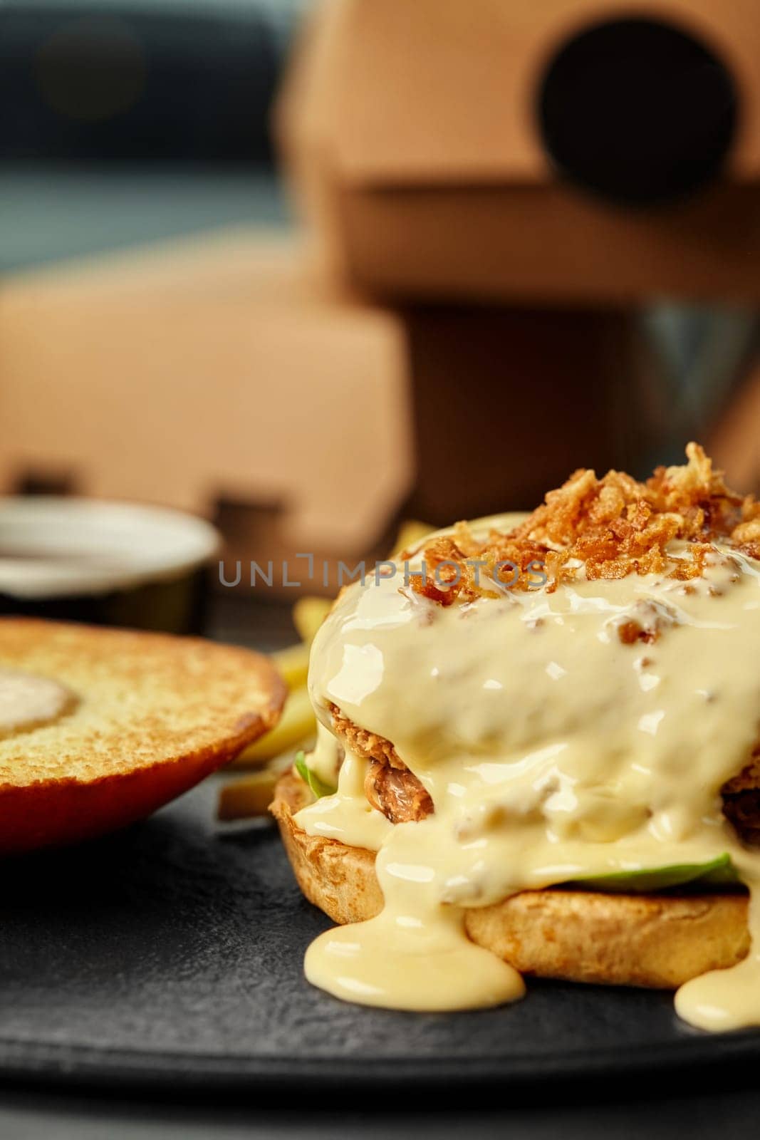 Close-up of open burger with breaded patty smothered in creamy cheese sauce sprinkled with crispy fried onions, with toasted top bun placed to side, served on slate plate