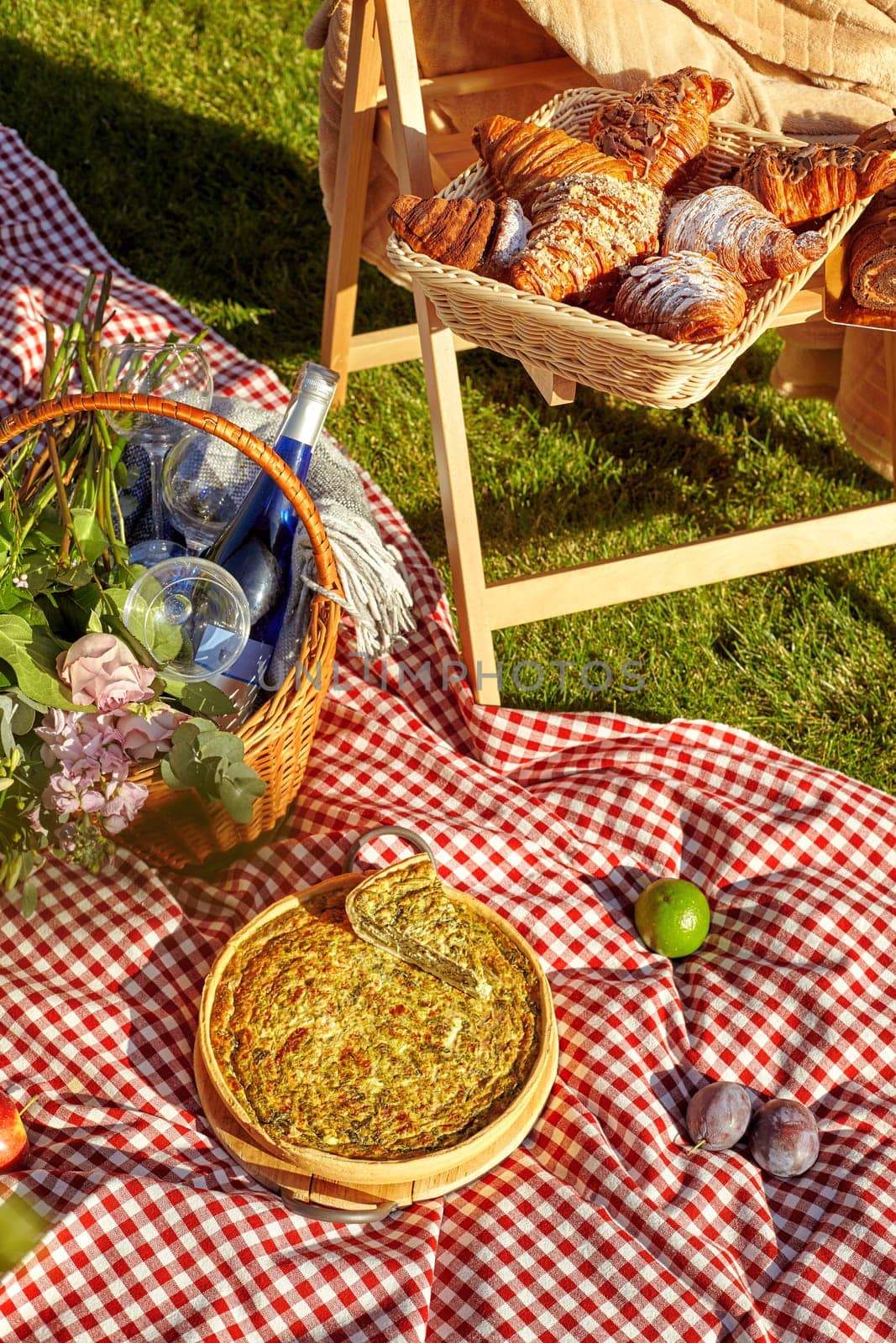 Appetizing savory quiche and sweet puff croissants with fresh fruits and bottle of wine arranged on wooden chair and red and white checkered blanket on lush green grass during summer picnic.