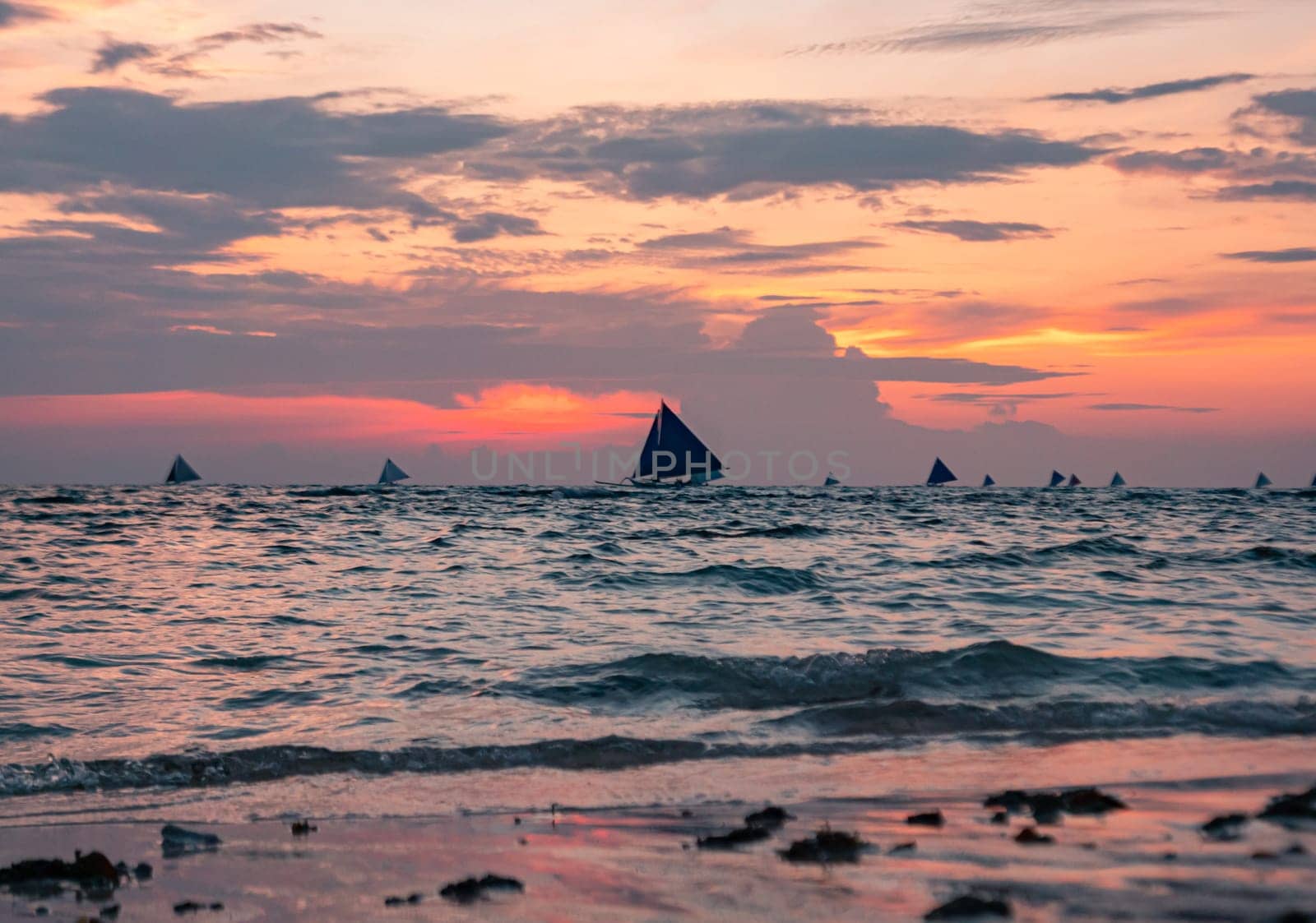 As the sun sets, the tranquil beach is bathed in soft hues of pink and purple. Boats with sails are visible on the horizon, gently drifting on the water. Boracay, Philippines.