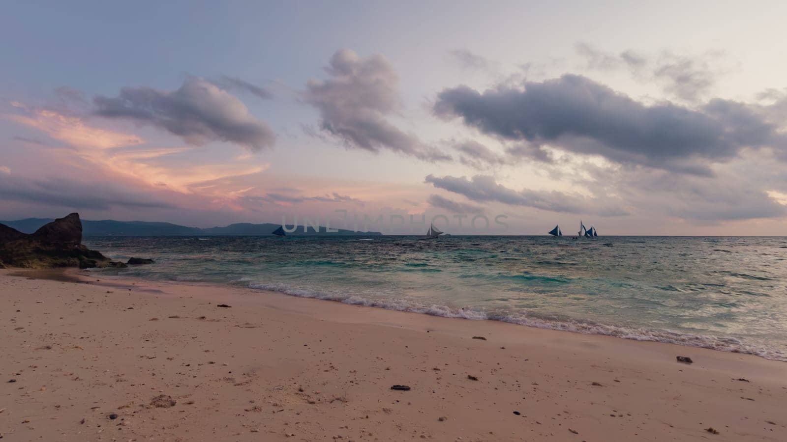 As the sun sets, the tranquil beach is bathed in soft hues of pink and purple. Boats with sails are visible on the horizon, gently drifting on the water. Boracay, Philippines.