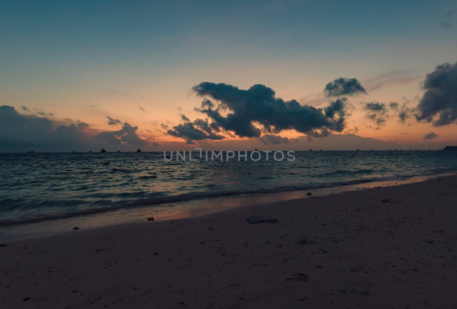 As the sun sets, the tranquil beach is bathed in soft hues of pink and purple. Boats with sails are visible on the horizon, gently drifting on the water. Boracay, Philippines.