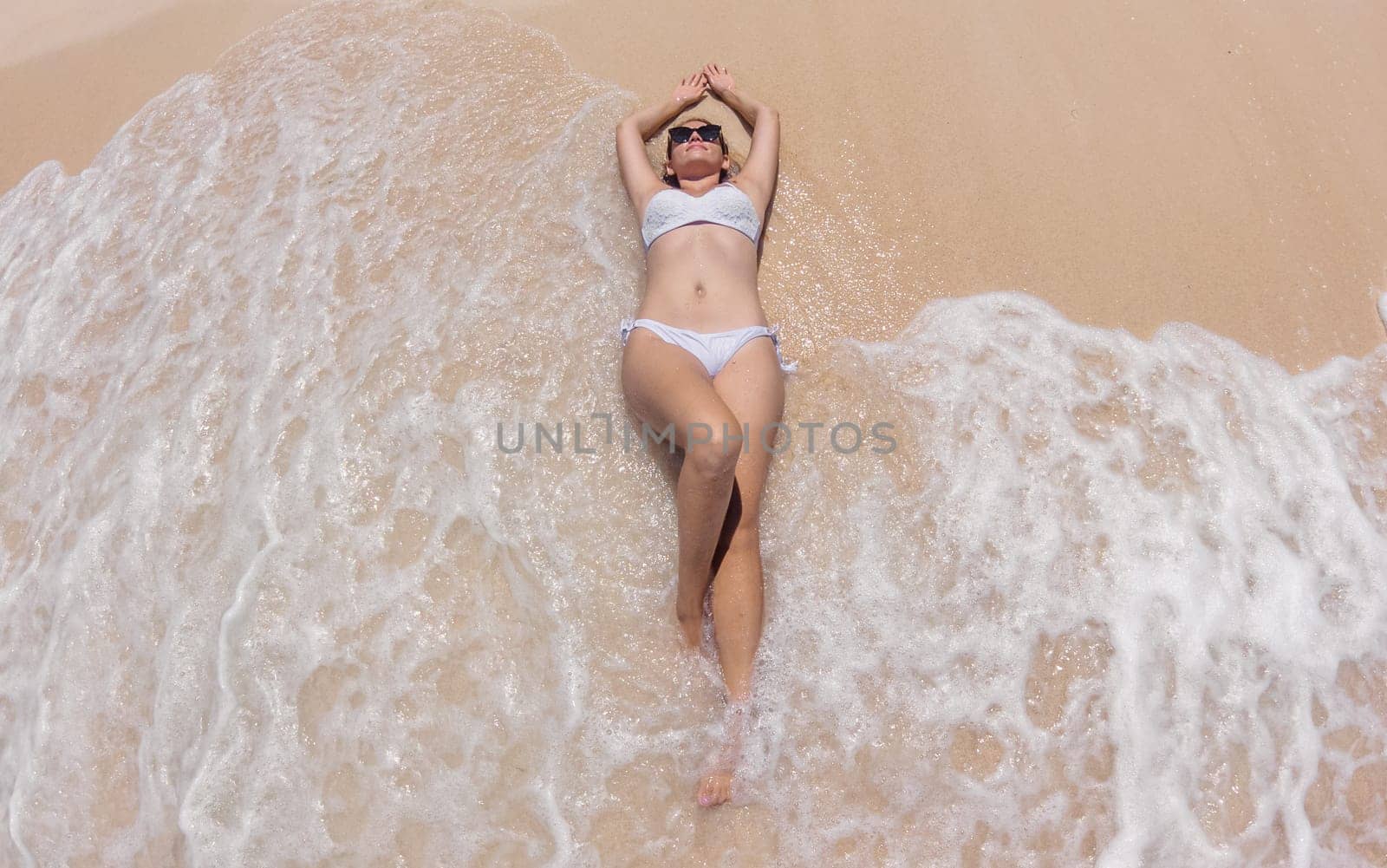 A woman lies on a sandy beach, her arms resting behind her head, as gentle waves lap around her. She is wearing a white bikini and sunglasses, enjoying the sunny afternoon at the shoreline.