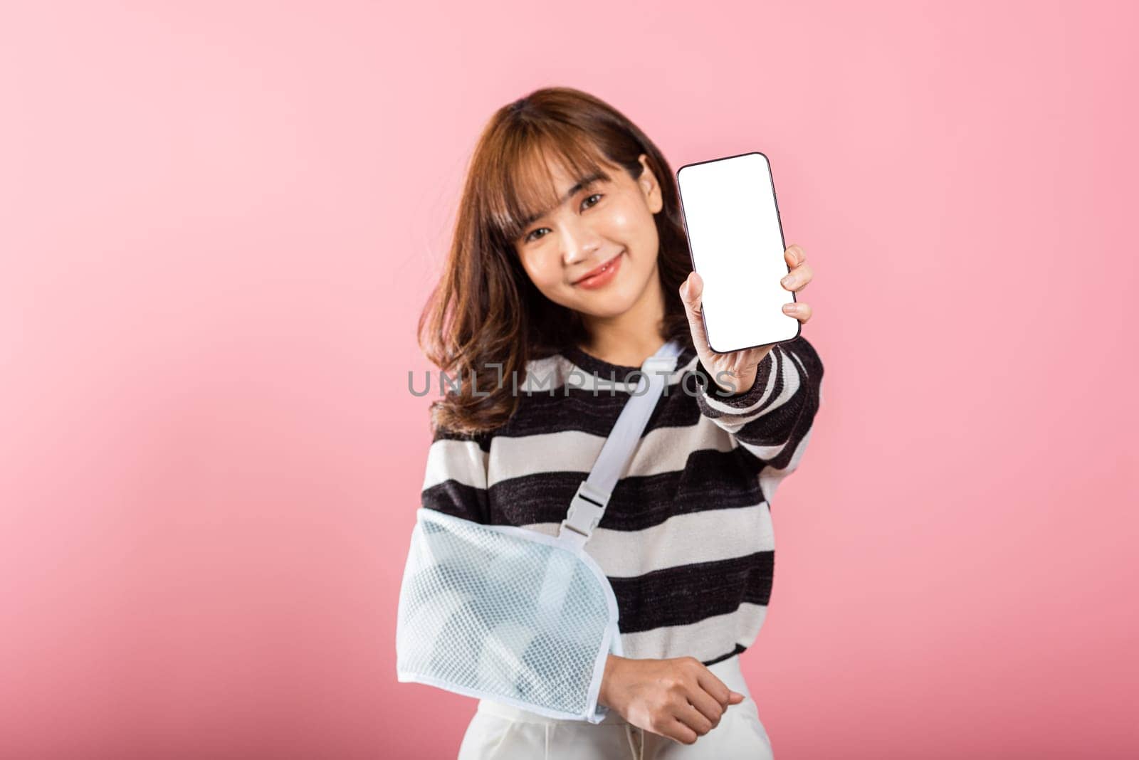 Happy Asian woman with a broken arm, wearing an arm splint, smiles while displaying her smartphone screen. Coping with pain and recovery, isolated on pink.
