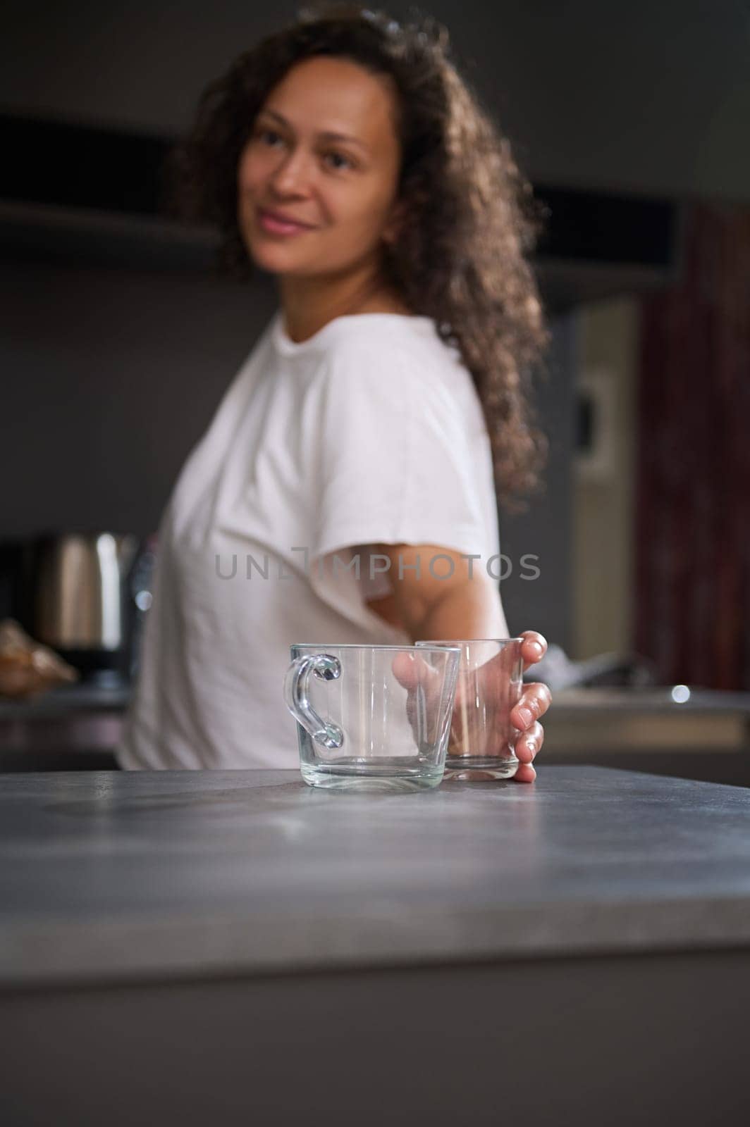 Happy woman taking out a glass cup from a cupboard, putting it on the kitchen counter, smiling looking aside, standing in modern minimalist home kitchen interior. Morning routine. Healthy lifestyle