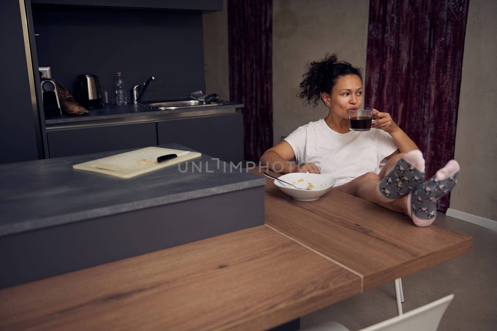 Pretty woman in pajamas, relaxing over cup of coffee. Happy young adult sitting at kitchen table, drinking hot coffee in the morning during her breakfast. Full size portrait
