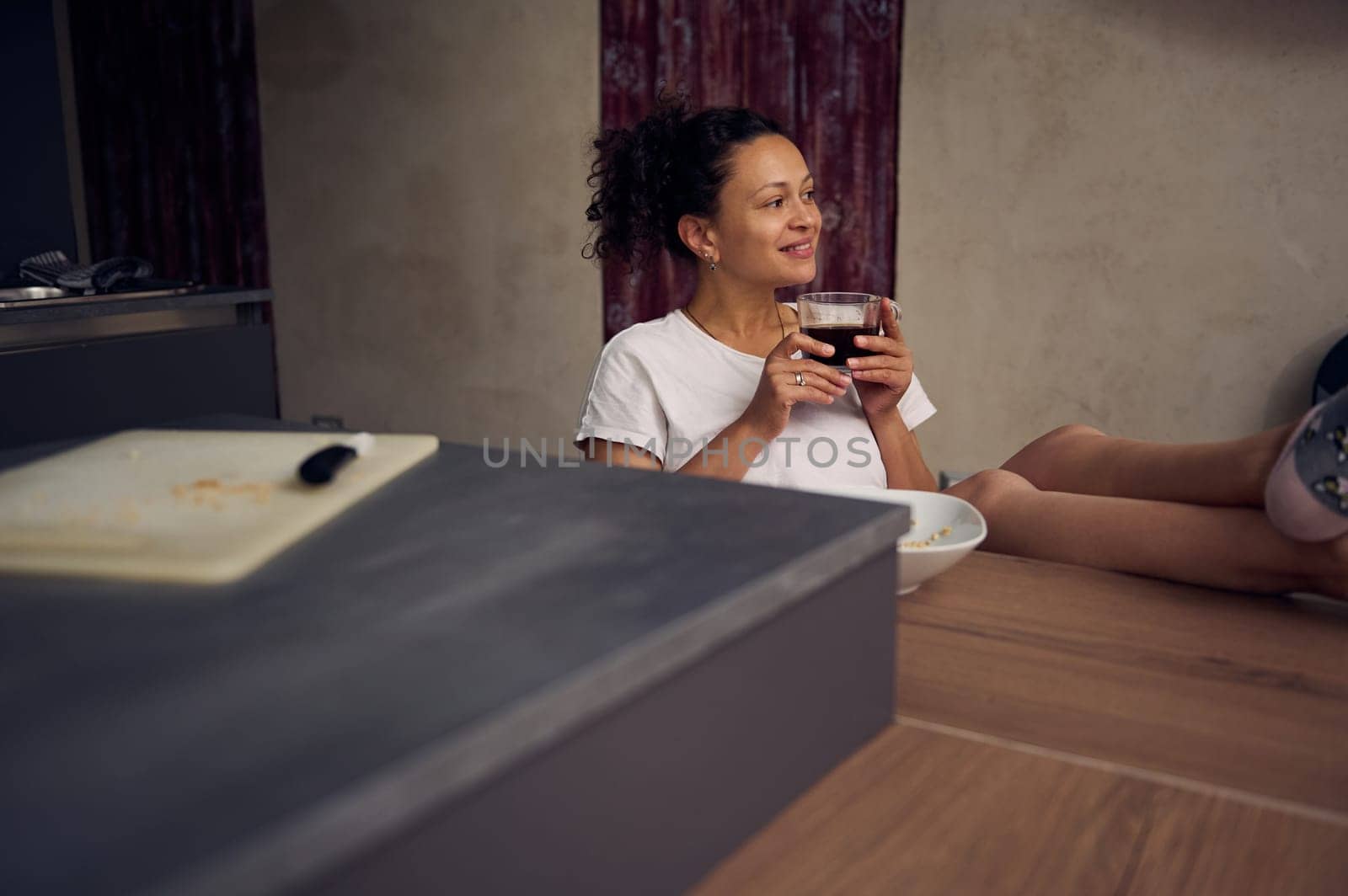 Middle aged pretty woman in pajamas, smiling looking away, relaxing over cup of coffee. Happy young adult sitting at kitchen table, drinking hot coffee in the morning during her breakfast