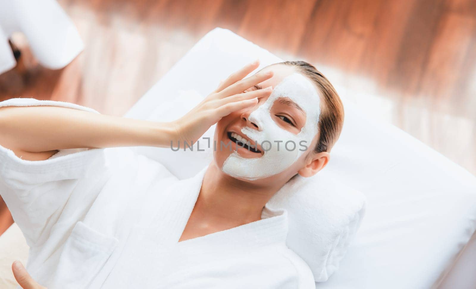 Serene daylight ambiance of spa salon, top view woman lying on massage table smiling and rejuvenating with face cream spa massage. Facial skin spa treatment and beauty care concept. Quiescent