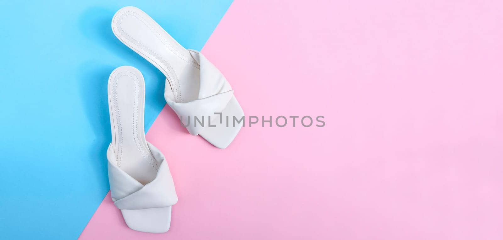 One pair of white female heeled flip flops lie to the left against a blue-pink background with copy space to the right of the flat lay close-up. The concept of fashion, beauty, shoes.