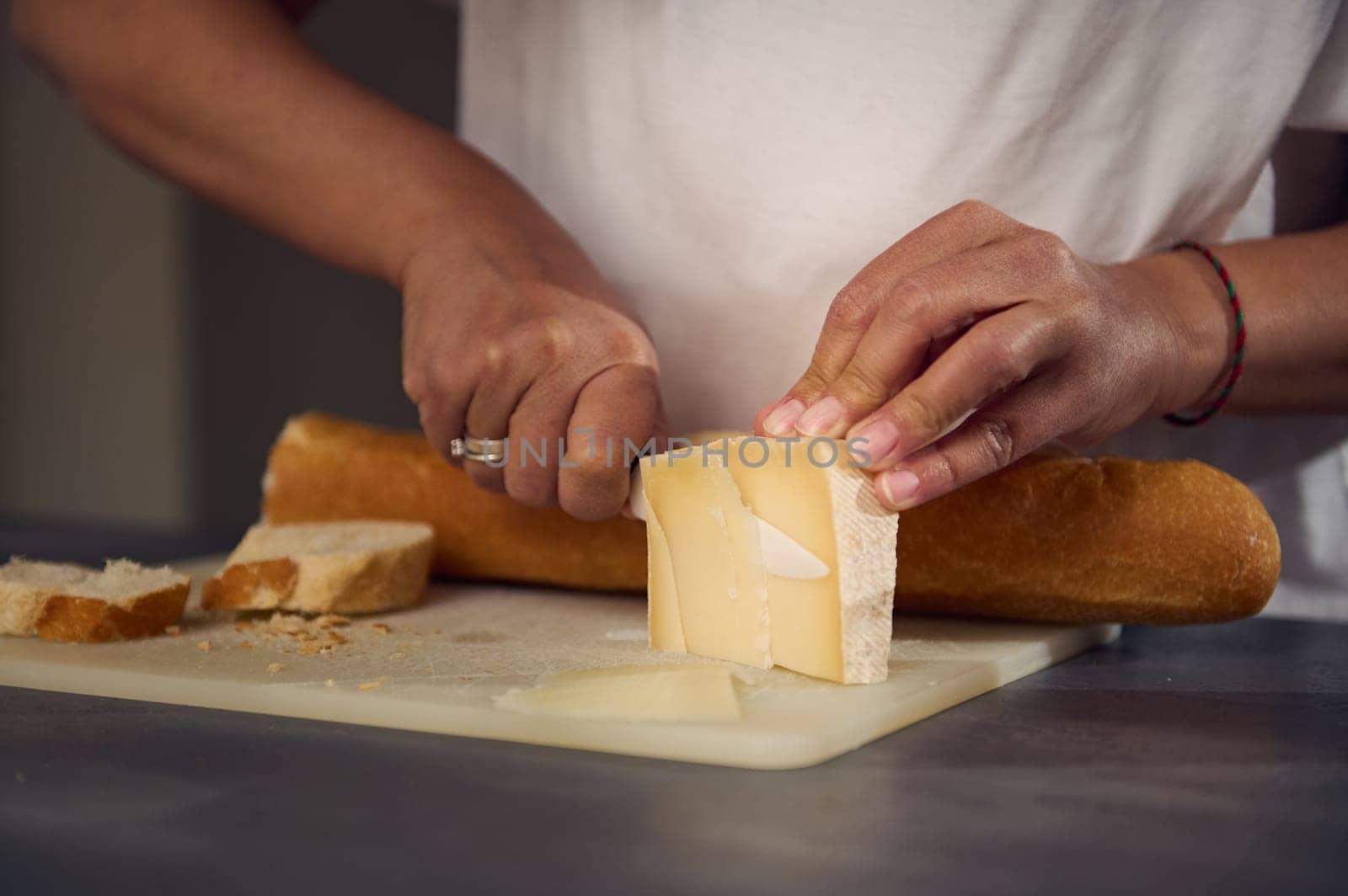 Close-up hands of a woman slicing cheese on cutting board, using a kitchen knife, preparing sandwiches with a loaf of whole grain wholesome integral bread baguette. Woman preparing breakfast