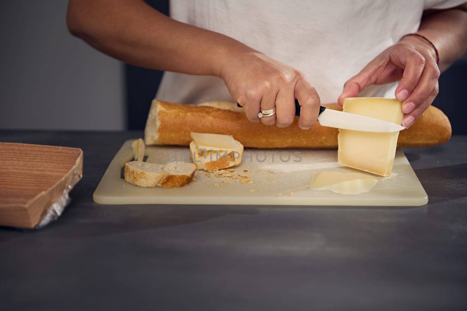 Close-up hands of a woman in white t-shirt, cutting cheese and bread, preparing sandwiches for snack, standing at kitchen countertop with fresh organic ingredients. Food and drink concept