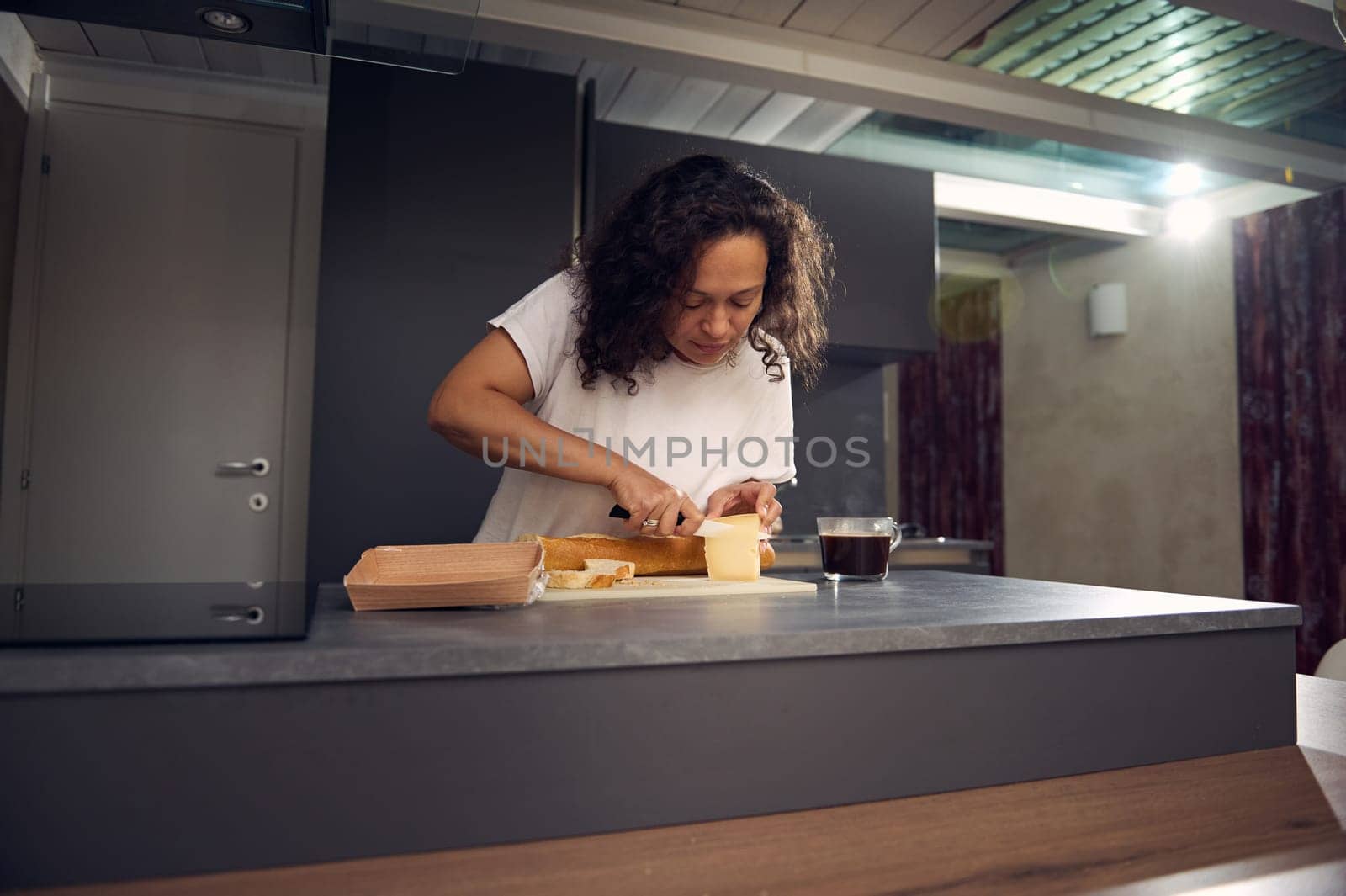 Curly haired multi ethnic adult woman slicing cheese on cutting board, using a kitchen knife, preparing sandwiches with a loaf of whole grain wholesome integral bread baguette for snack or breakfast