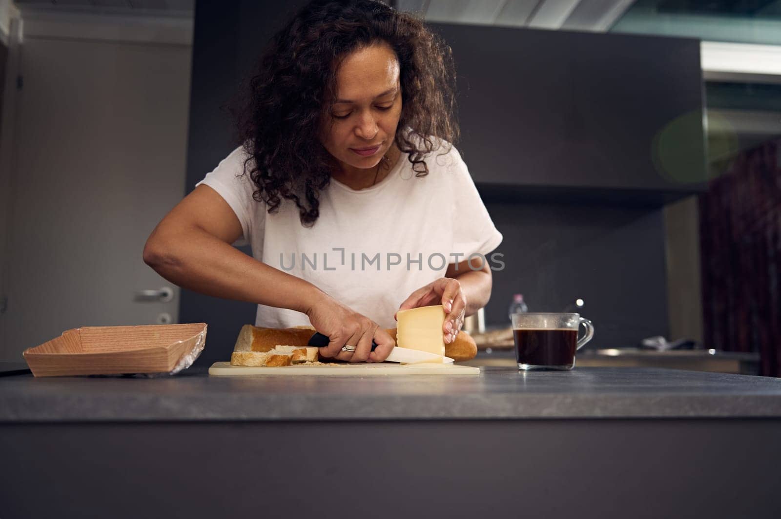 Beautiful multi ethnic young woman in white t-shirt, putting a slice of cheese over piece of bread, preparing sandwiches for snack