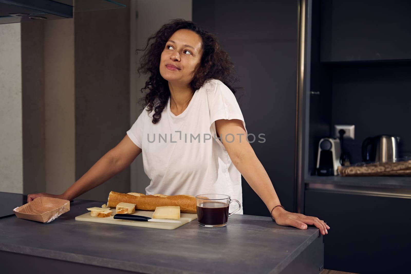 Multi ethnic pensive happy woman smiling looking away, standing at kitchen counter with a cup of freshly brewed coffee and fresh sandwiches for breakfast