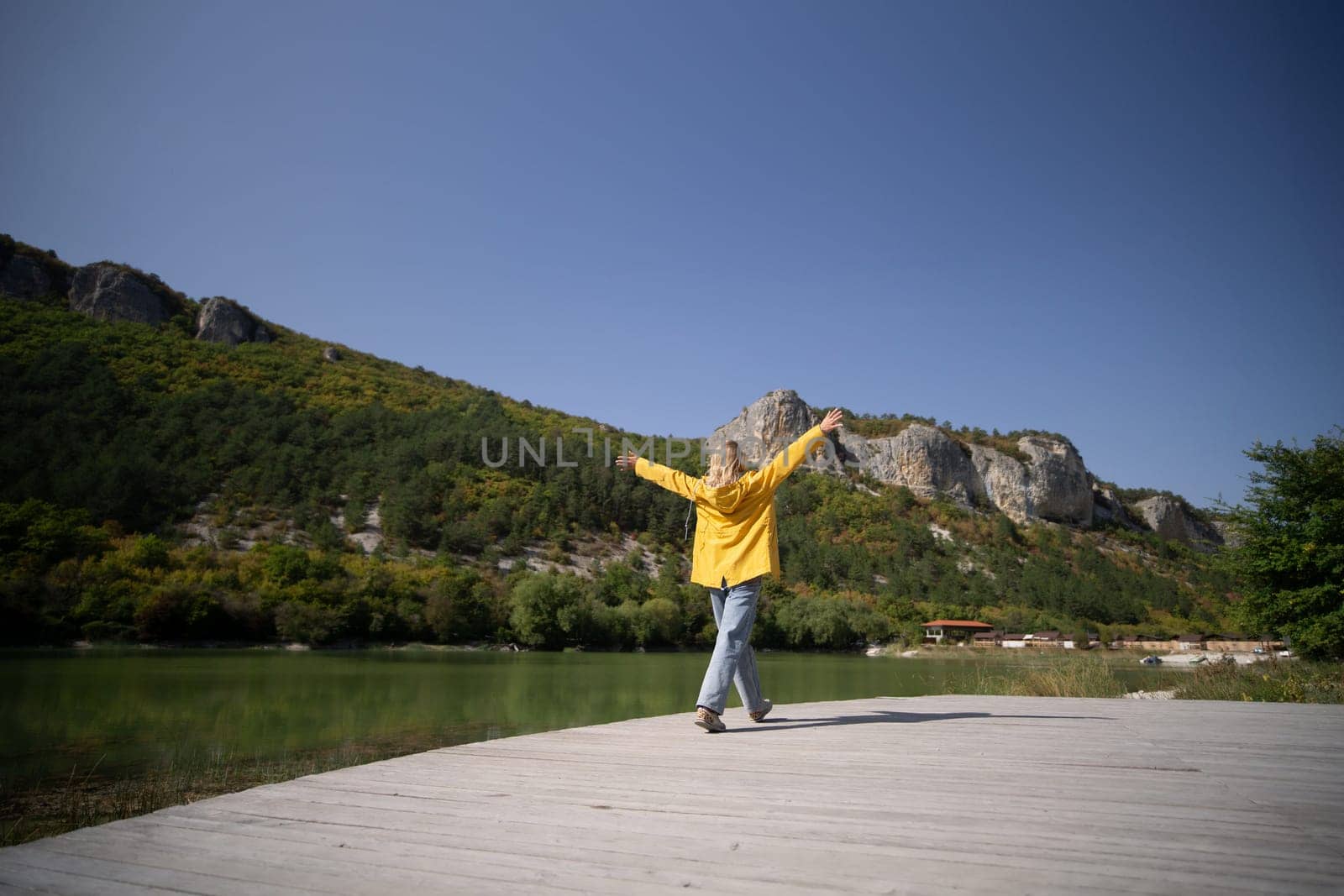 A woman in a yellow jacket is walking on a wooden boardwalk near a lake. The sky is clear and the mountains in the background create a serene and peaceful atmosphere