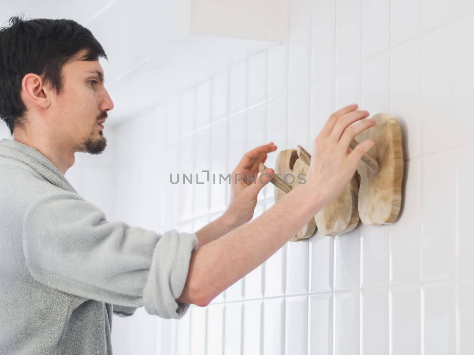 A young handsome caucasian guy in a gray fleece bathrobe with rolled up sleeves hangs a new wooden stylish and eco-friendly hanger on a wall with white tiles in the bathroom, close-up from below. The concept of installing a hanger, installation, dismantling, repair, cleaning the house, bathroom, eco-friendly accessory.