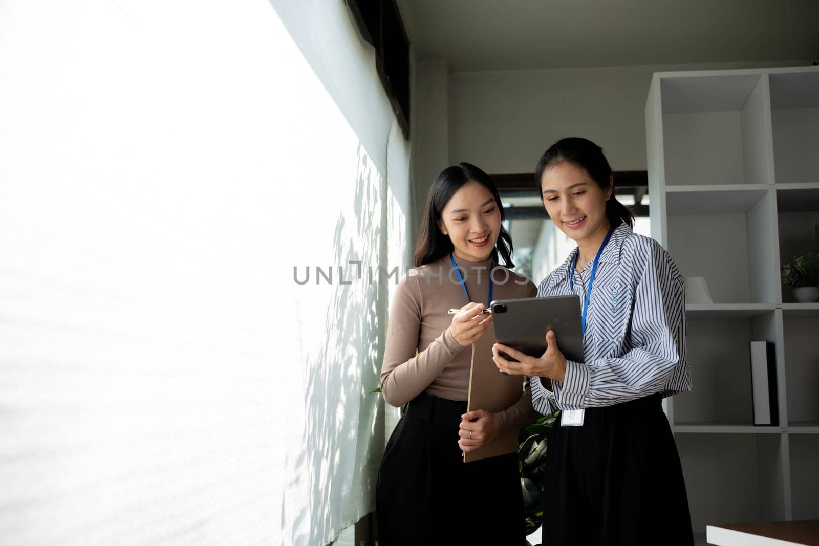 Two Asian businesswomen discussing work using a tablet in a modern office. Concept of teamwork and professional collaboration.