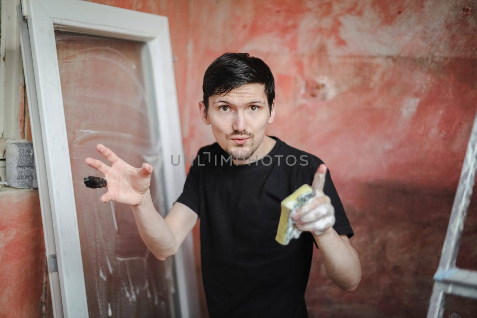 Handsome caucasian young brunette man emotionally speaks to the camera waving his hands with a bandaged finger with a black adhesive tape and a soapy sponge against the background of a window frame and a red wall, close-up side view with selective focus. The concept of home renovation, washing window frames.