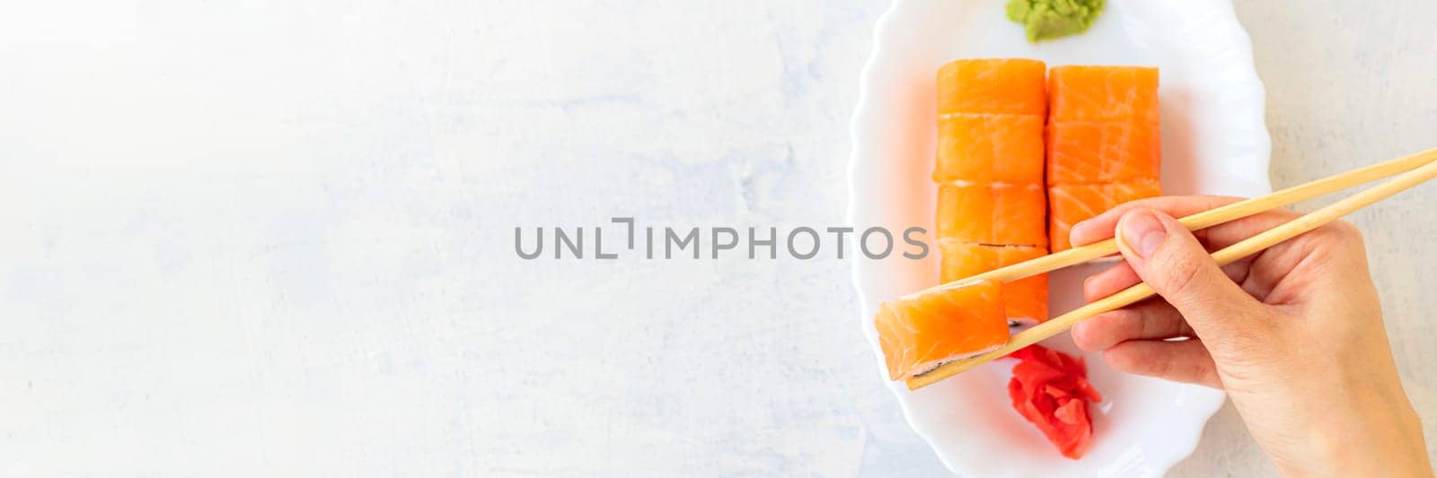 Banner of female hands holding Japanese sushi or Philadelphia roll with wooden chopsticks on a light textured background by Leoschka