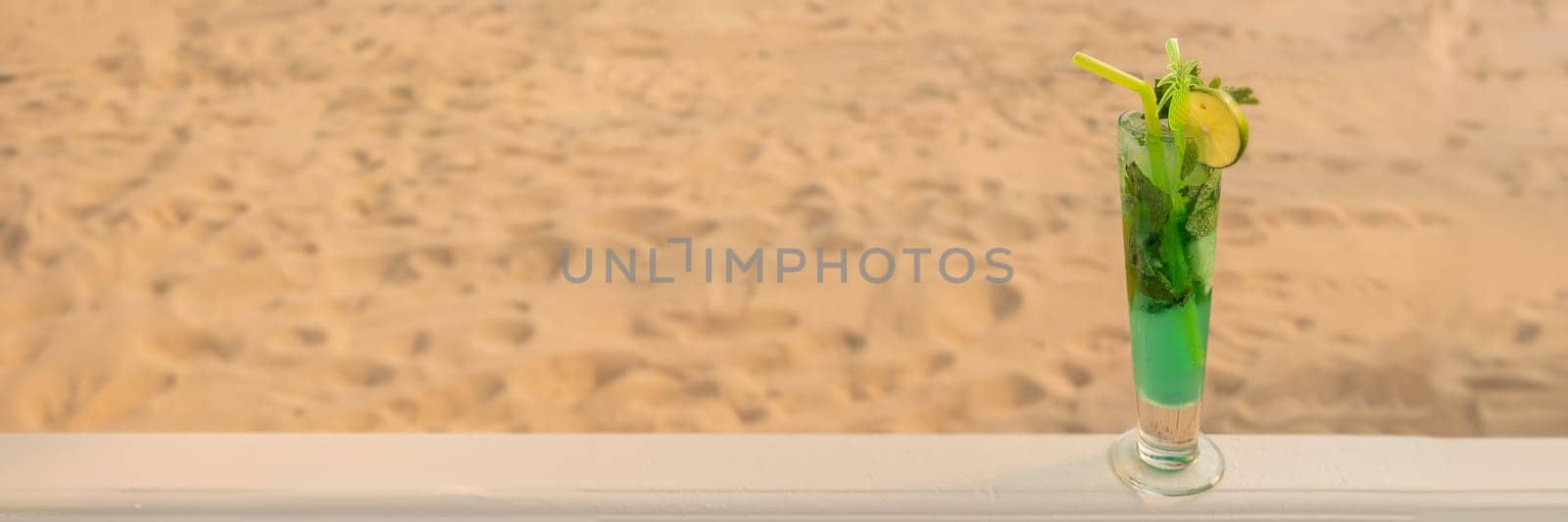 Banner of tropical green cocktail with straw, mint and lime on background of golden sand. the concept of relaxing and eating by the sea on a bright, hot and sunny day on the beach. soft focus. copy space