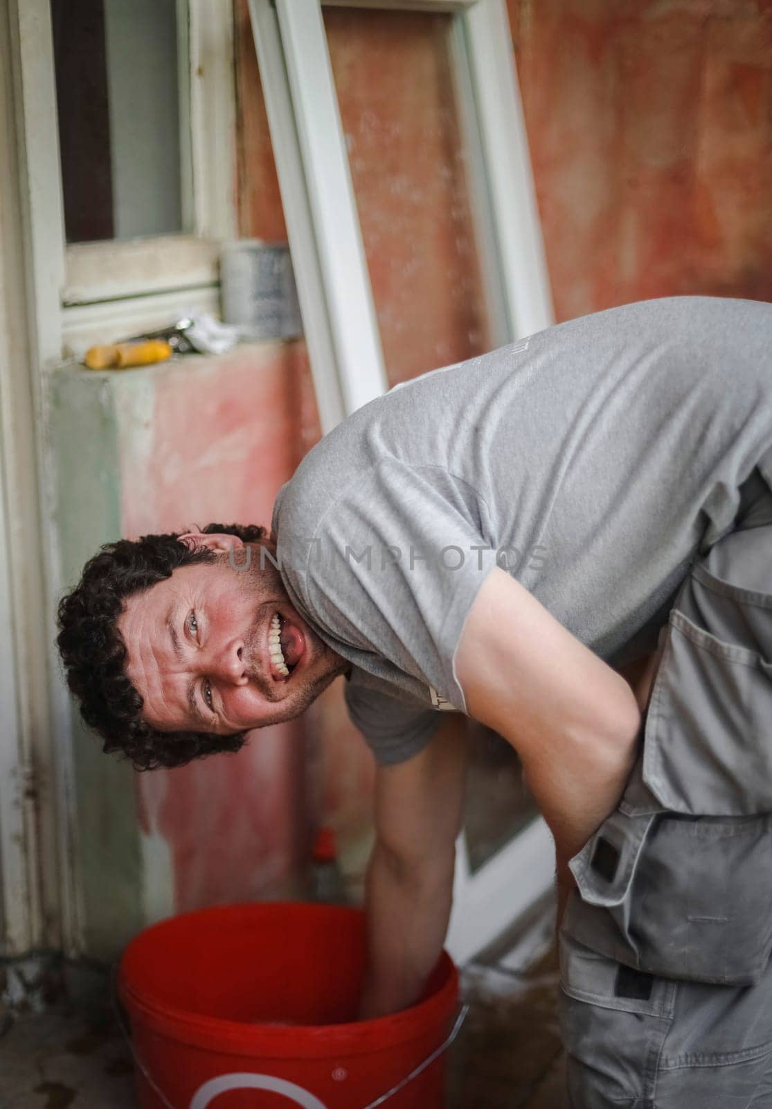 Handsome caucasian young brunette male with curly hair smiling showing tongue standing at an angle with his hand in a red bucket of water against the background of a window frame and a red wall, close-up side view with selective focus. The concept of home renovation, washing window frames.