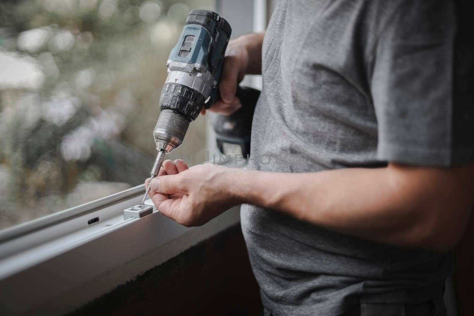 Young caucasian man holding drill and screw getting ready to install fittings on plastic window frame in renovation room, side view closeup with selective focus. The concept of home renovation, washing window frames.