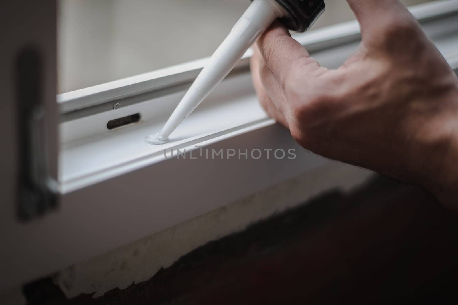 The hands of a young caucasian man squeezes white silicone into a small hole in the window frame, close-up side view with selective focus.