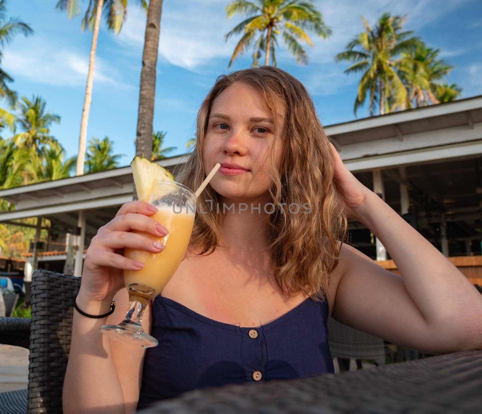 A young woman sips on a tropical beverage while sitting at an outdoor restaurant. Palm trees and a blue sky with scattered clouds are visible in the background.