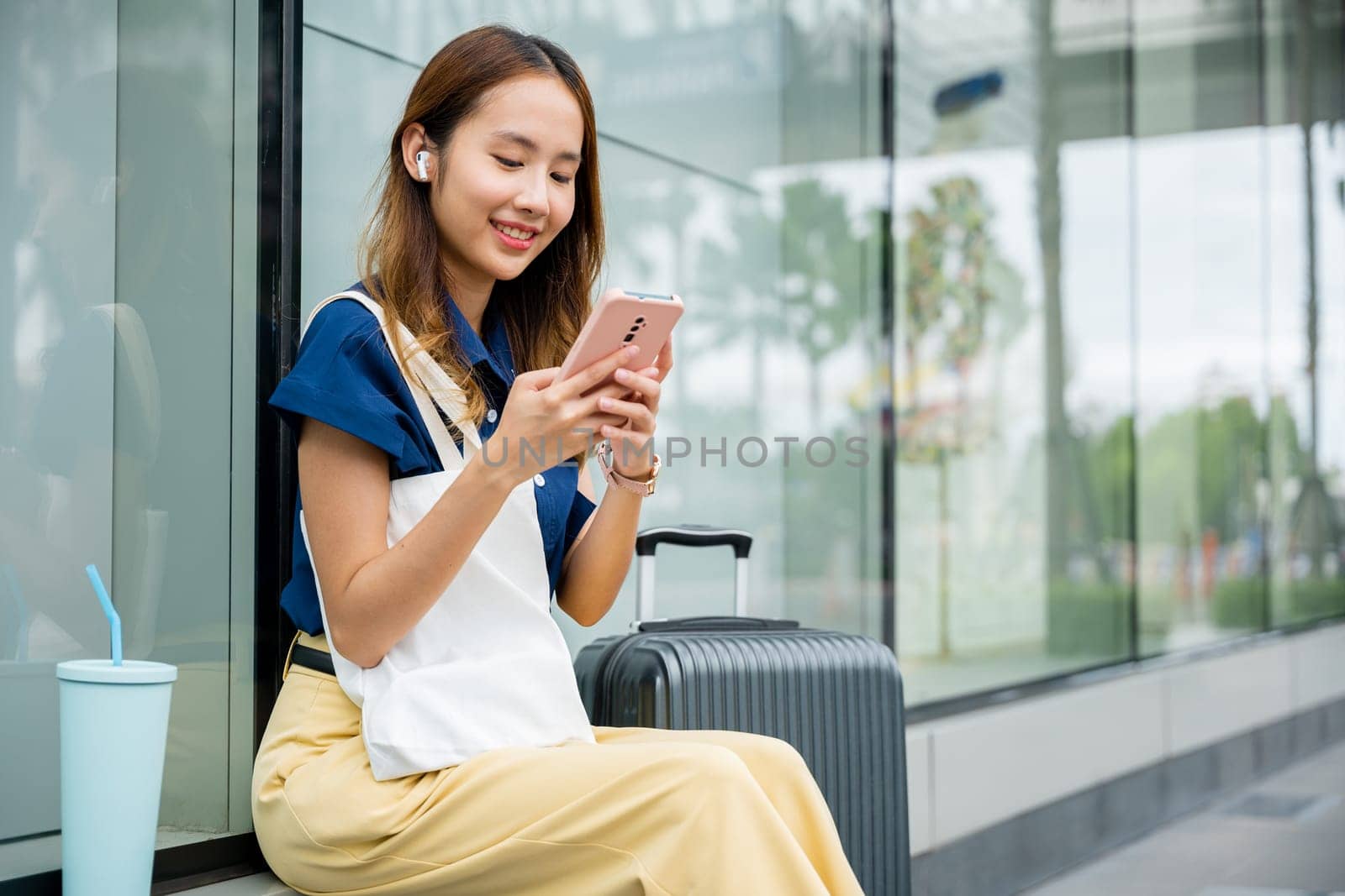 A woman sitting with her suitcase, using her mobile phone to send messages and stay connected while waiting for transportation in the city. She is modern and connected.