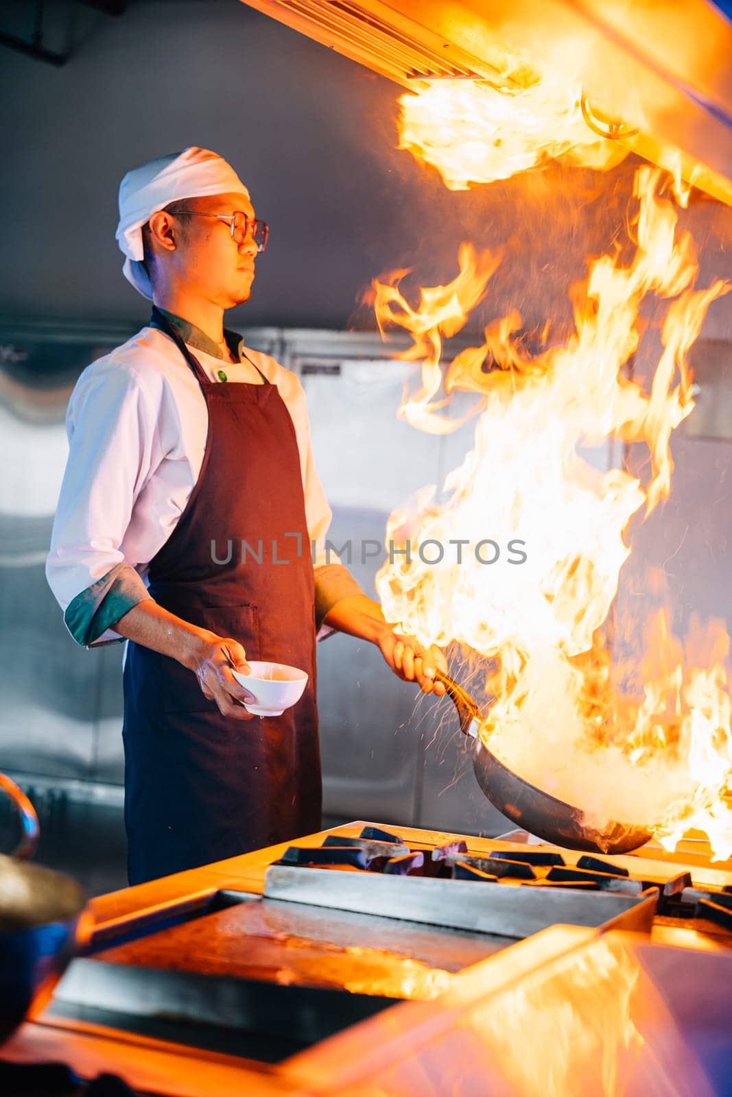 Closeup of chef hands in professional kitchen handling flaming wok. Expertise at work flames cooking food. Skilled chef busy in modern kitchen with flames. cook food with fire by Sorapop