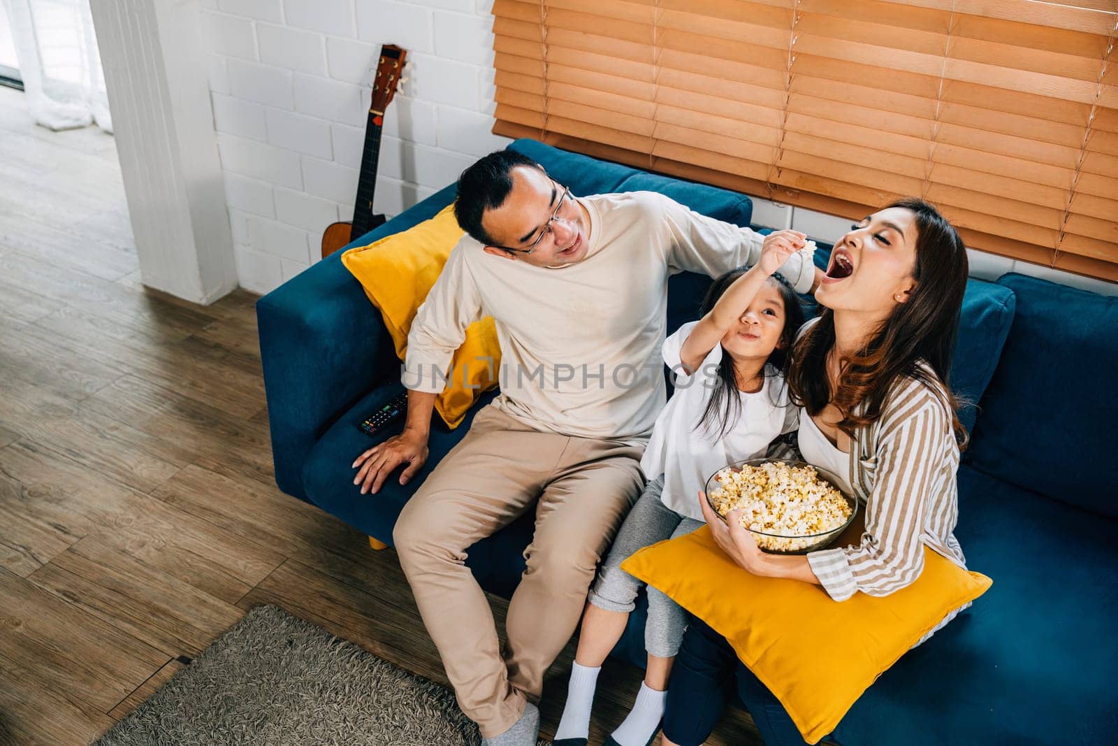 A happy family filled with joy watches TV while eating popcorn on modern sofa at home by Sorapop