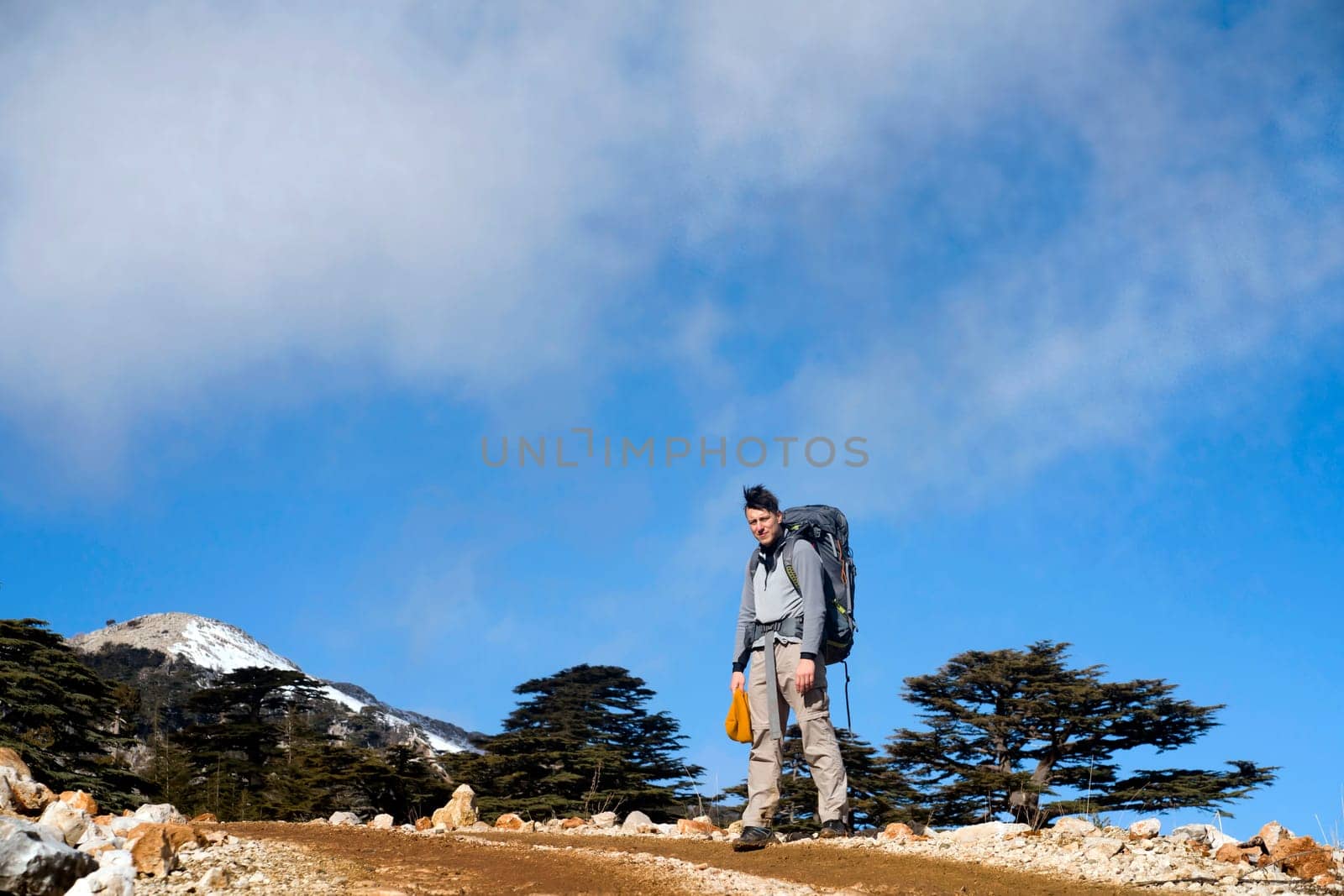 Young man travels, walks along a trail among the mountains with tourist equipment, the traveler stands against the background of a bright blue sky on the way to the top of the hill.