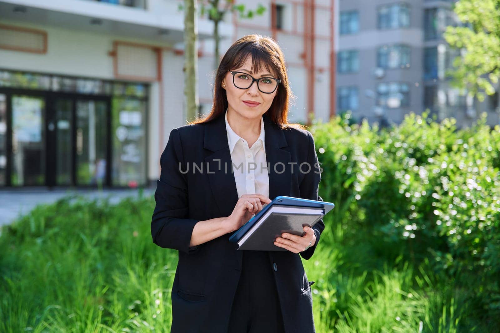 Middle-aged business woman in suit with digital tablet, outdoor building. Female teacher posing near school educational center, office worker entrepreneur accountant economist banker financier lawyer