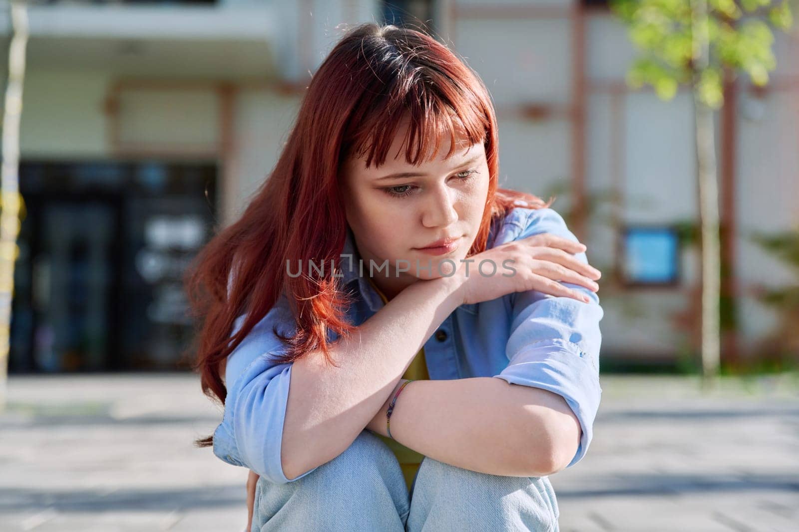 Upset sad unhappy young female sitting on steps. Pensive serious beautiful red-haired girl university college student sitting outdoor. Problems, difficulties, depression, mental health of young people