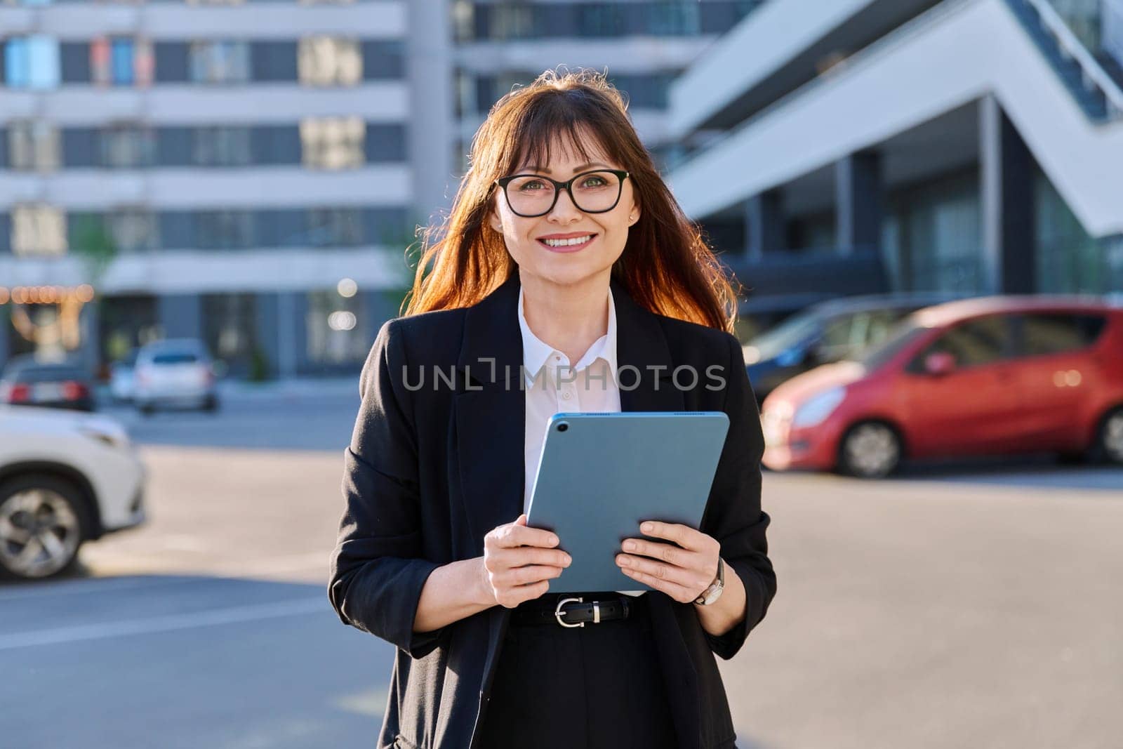 Mature confident successful business woman with digital tablet in black suit looking at camera outdoors, backdrop of modern city. Business, entrepreneurship mentoring insurance sales advertising work