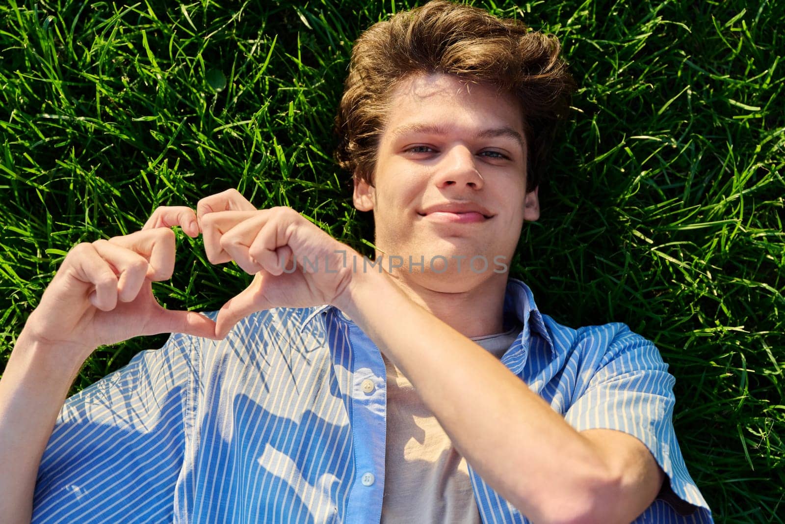 Top view, happy handsome smiling young male looking at camera, lying on background of green lawn grass, hand showing heart gesture