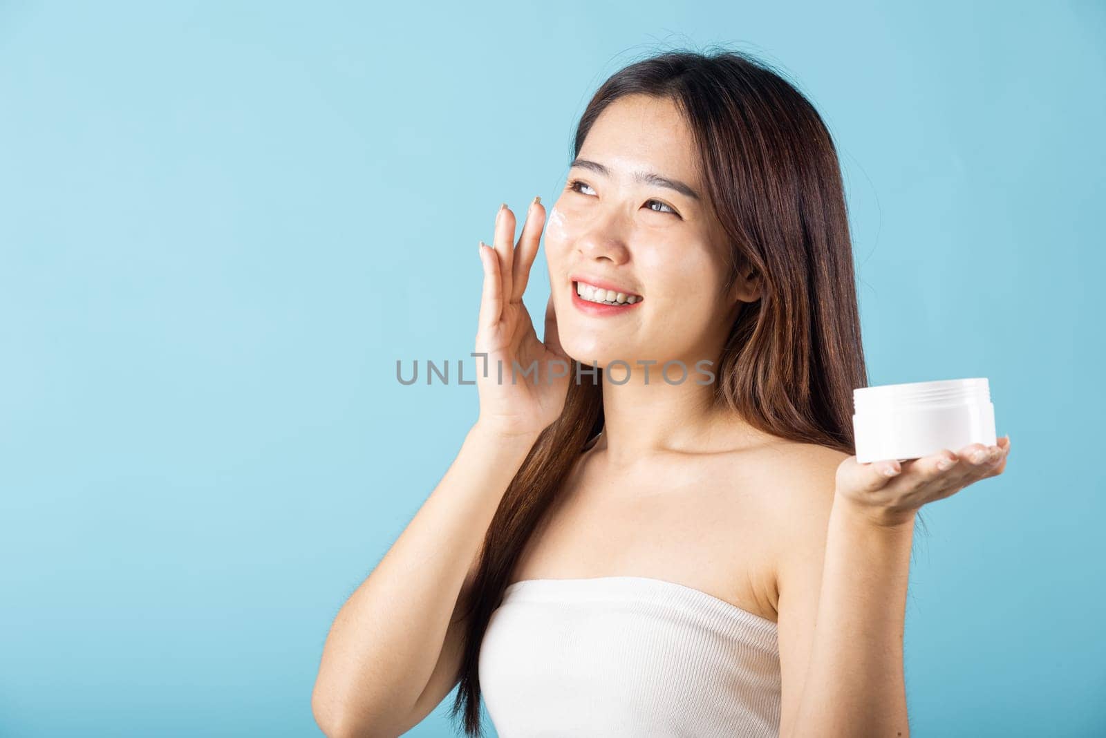 Asian young woman are not very beautiful hold jar of skin cream for face and apply anti-aging moisturizer studio isolated on blue background, Happy female pointing finger her face, pursuit of beauty