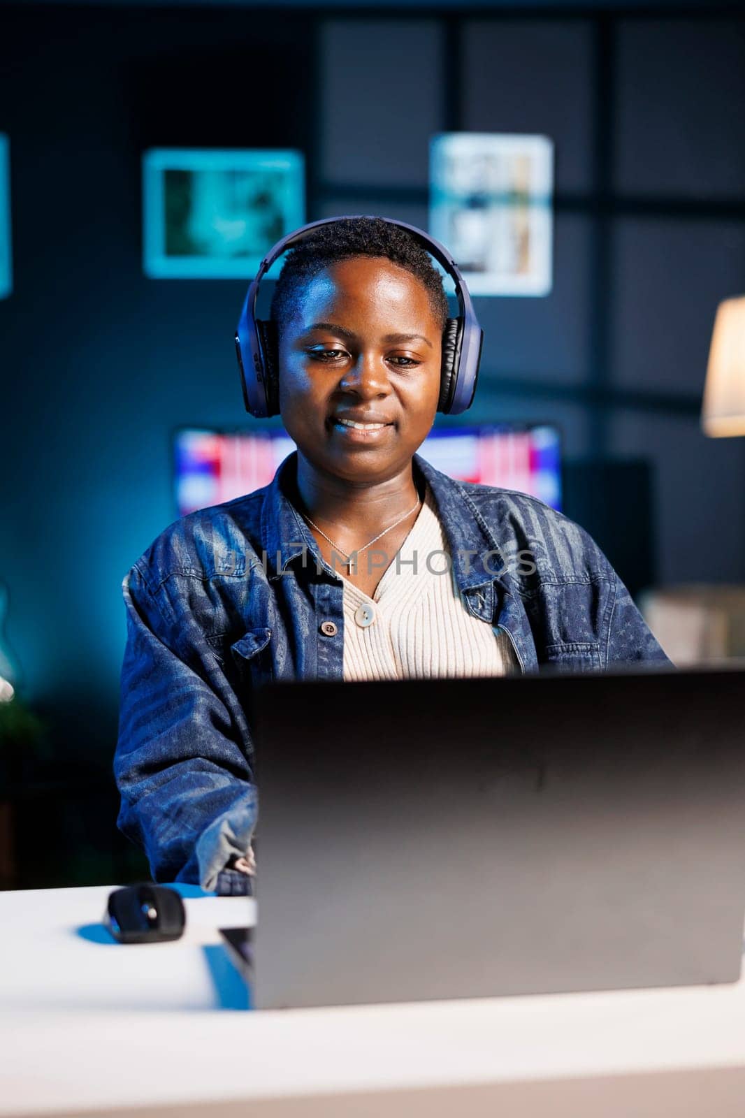 Freelancer working on a laptop, completing online tasks while listening to music through an audio headset. Portrait of African American woman using personal computer and wireless headphones.