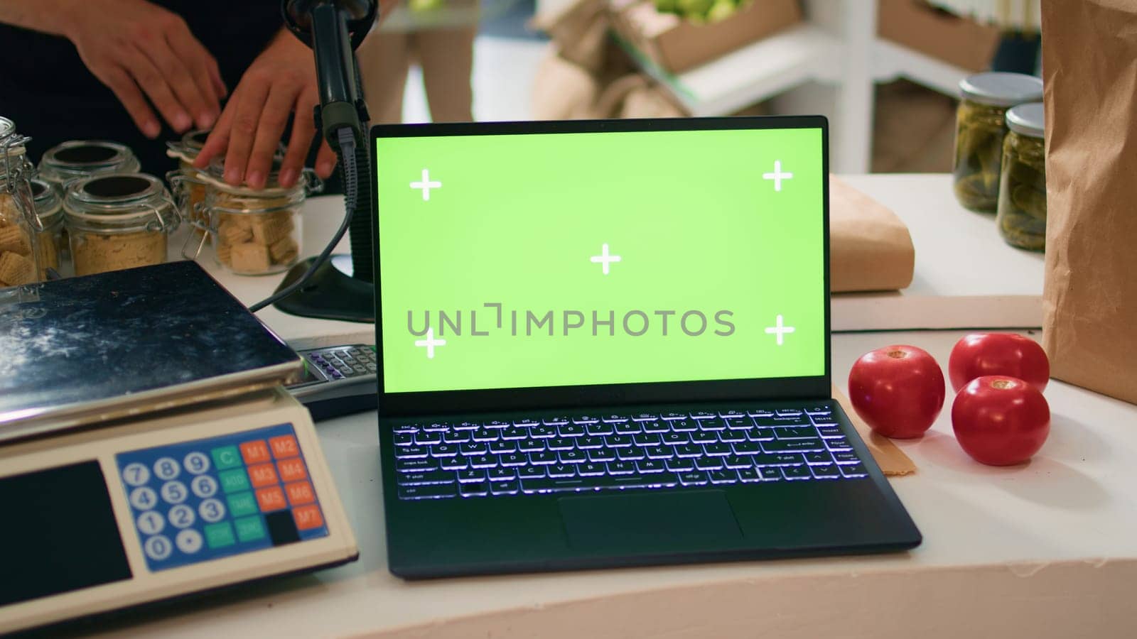 Vendor at register uses laptop running greenscreen display on device, young adult working at local organic supermarket looks at isolated copyspace. Seller waits for buyers at counter.