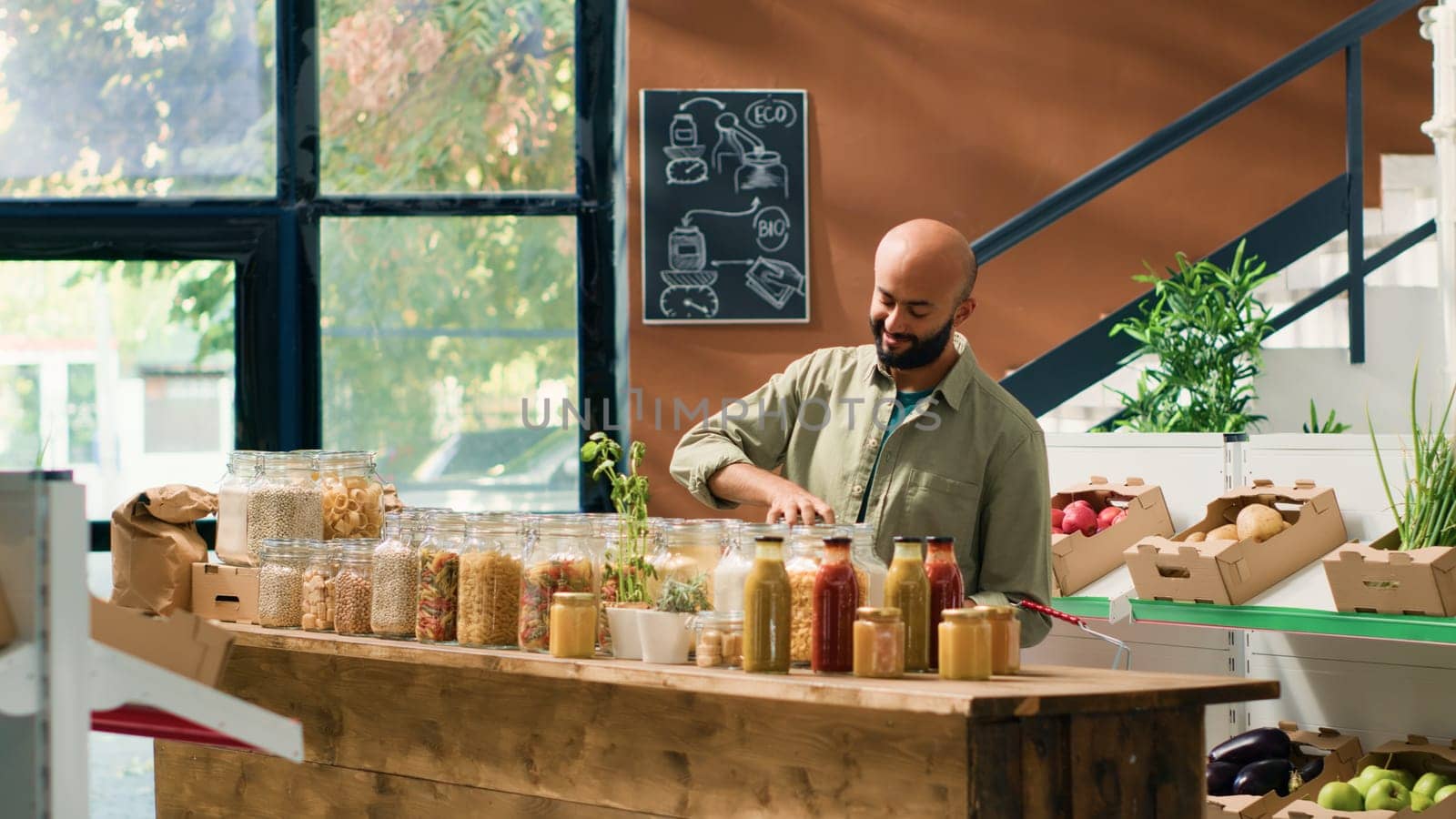 Middle eastern man in ecological food store examining organic homegrown fruits and vegetables, opening jars with sustainable pasta or spices. Customer admires fresh bio merchandise.