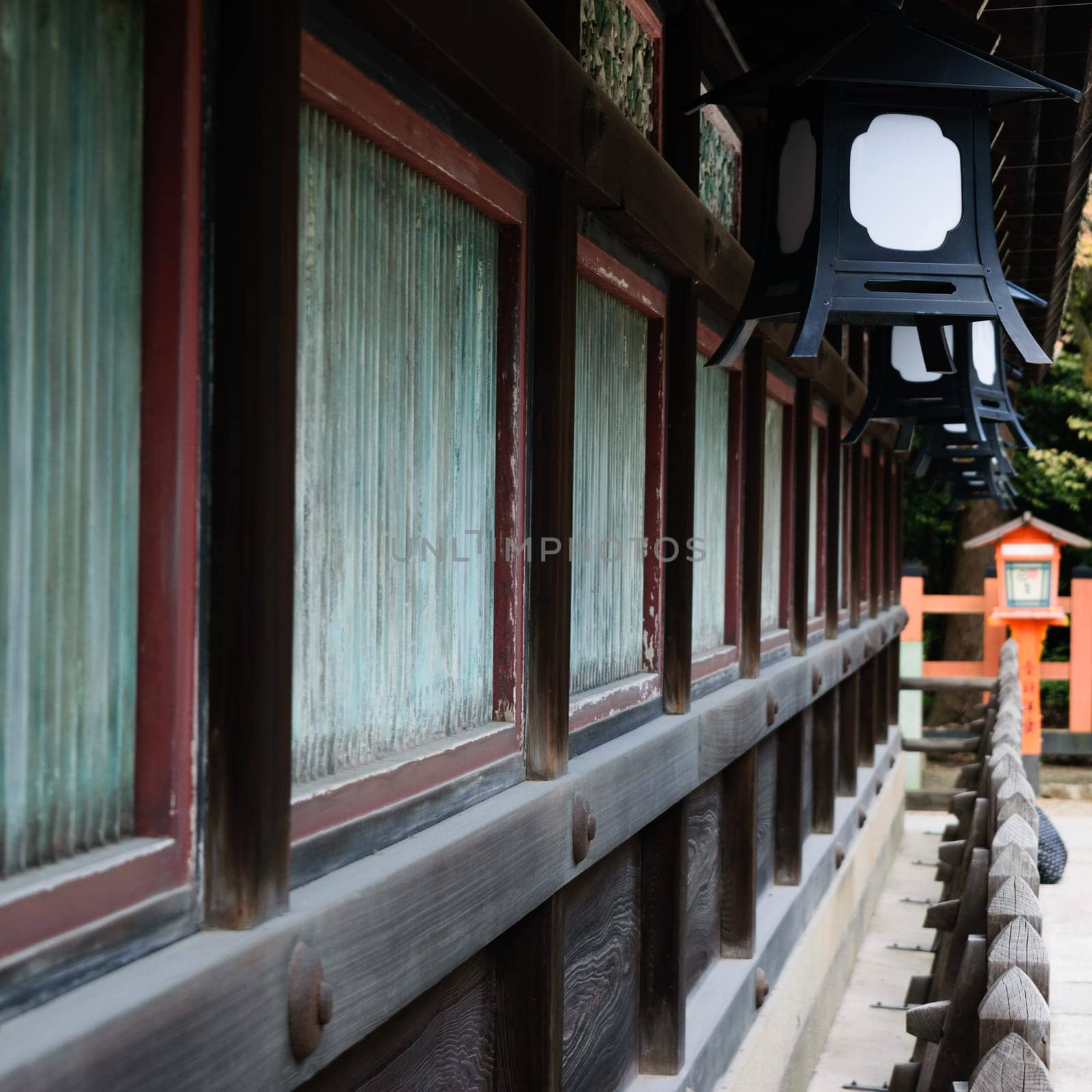 Close-up of a traditional Japanese building with wooden walls and metal lanterns. The building features green-tinted windows and a wooden fence in the foreground.