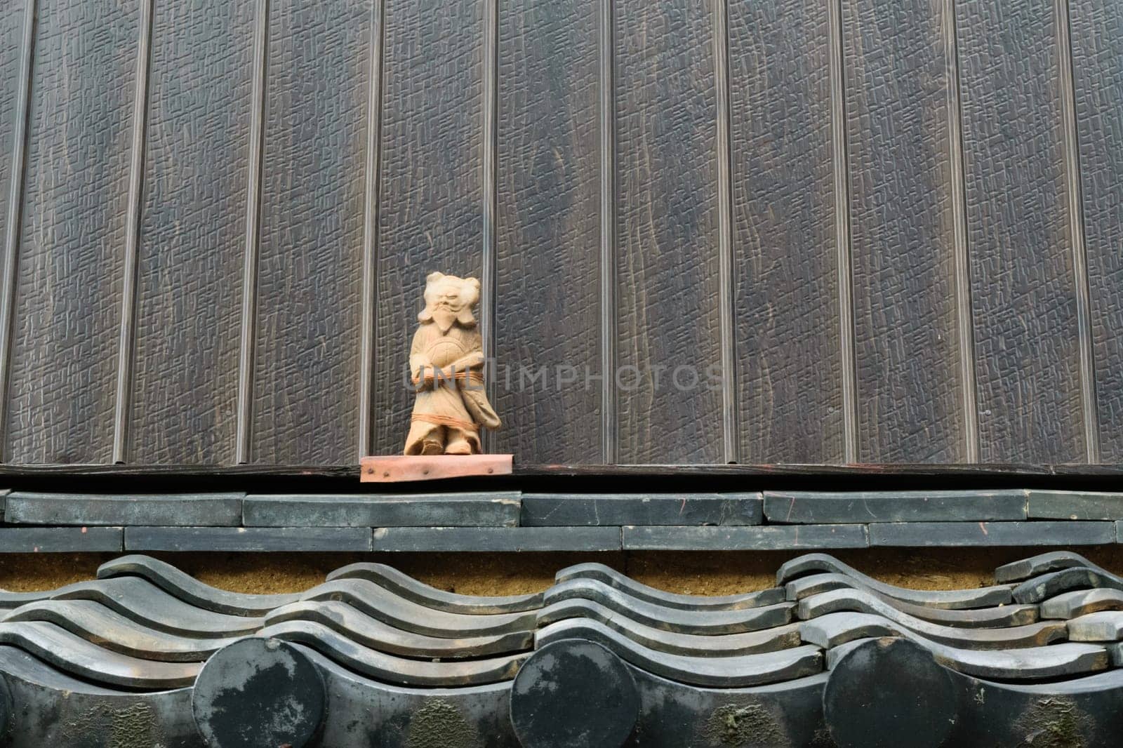 A small stone statue of a figure with a hat, standing on a traditional Japanese tiled roof against a textured wall.